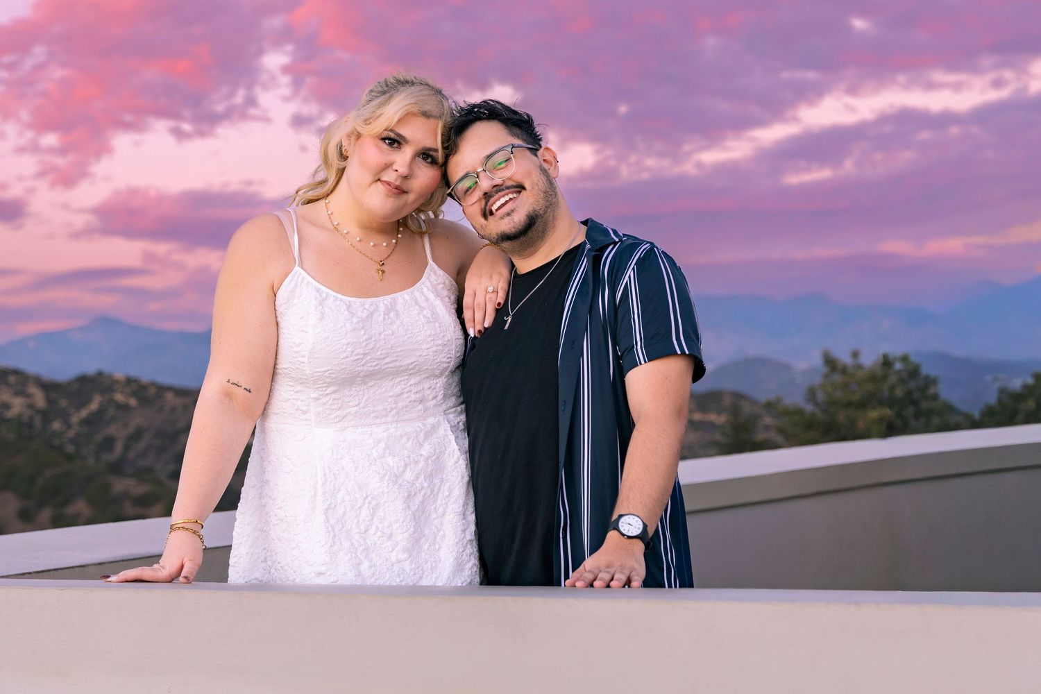 Two people share a romantic moment at sunset with mountains and pink sky in the background.