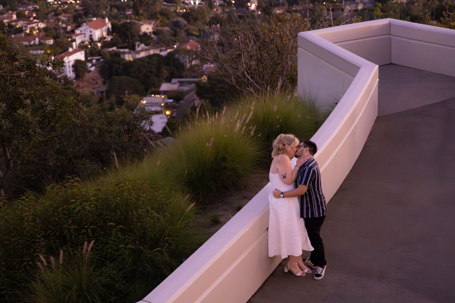 A romantic couple shares a tender moment at sunset on an outdoor balcony overlooking a city view.