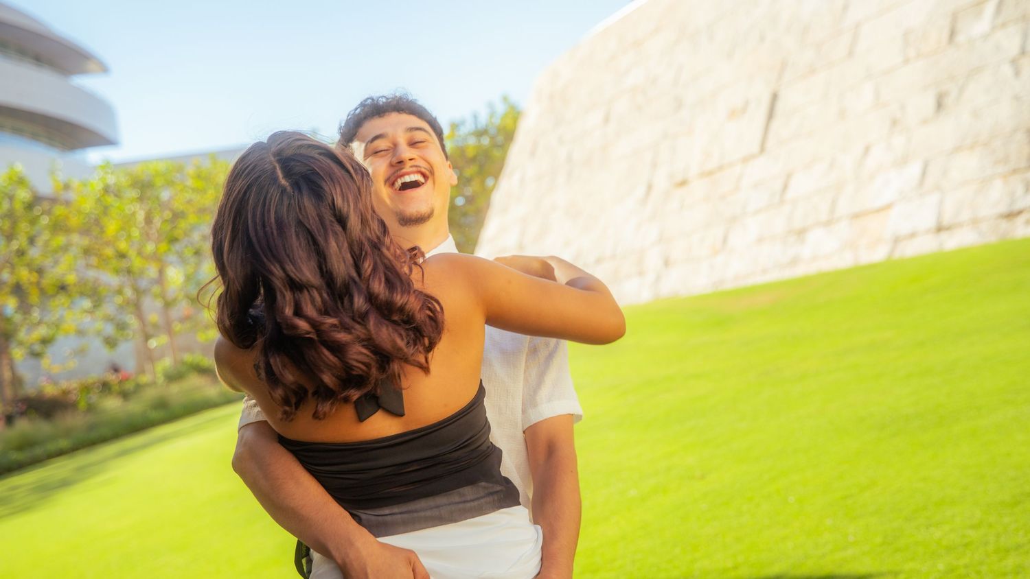 A joyful couple embraces and laughs together on a lush green lawn with a modern building in the background.