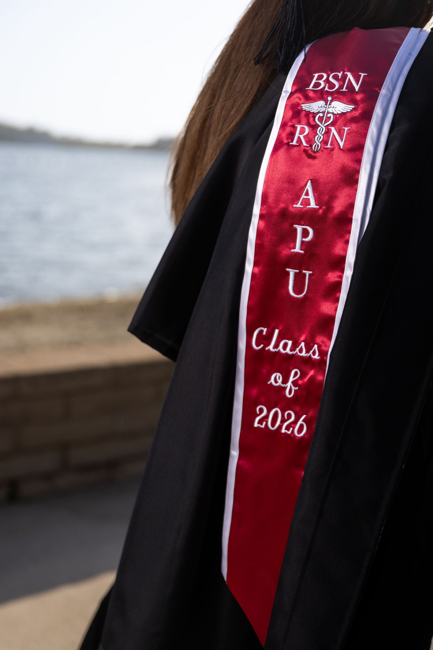 APU BSN RN Class of 2026 graduation sash on black gown near waterfront.