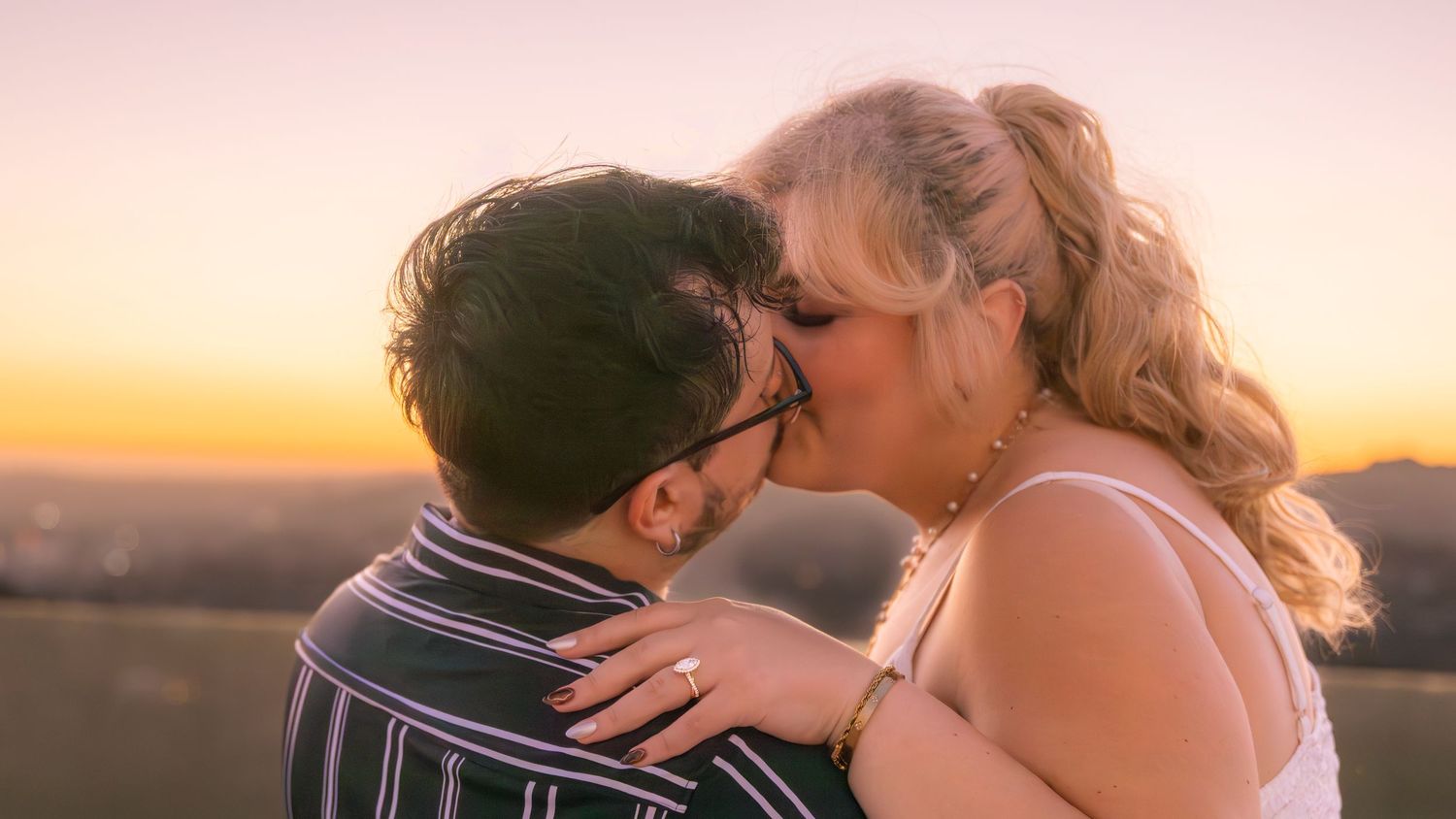A couple embraces in a romantic kiss at sunset against a warm golden sky.
