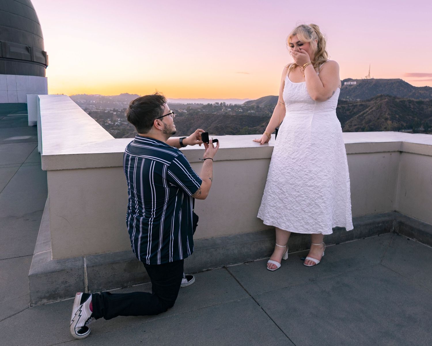 A couple shares a romantic moment during sunset on a rooftop balcony overlooking mountains.