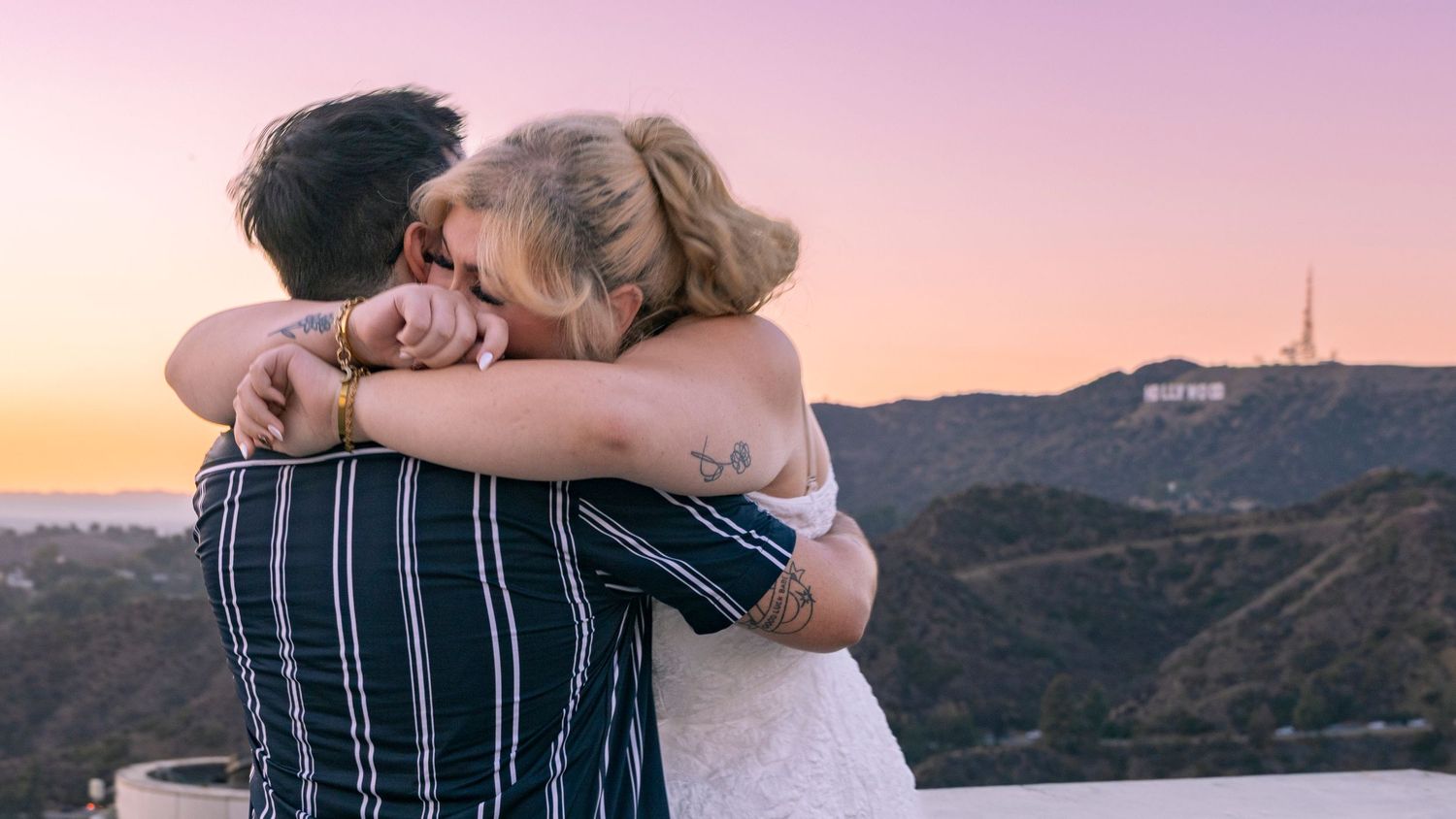 A couple embraces during sunset with mountains and coastline in the background creating a romantic silhouette.