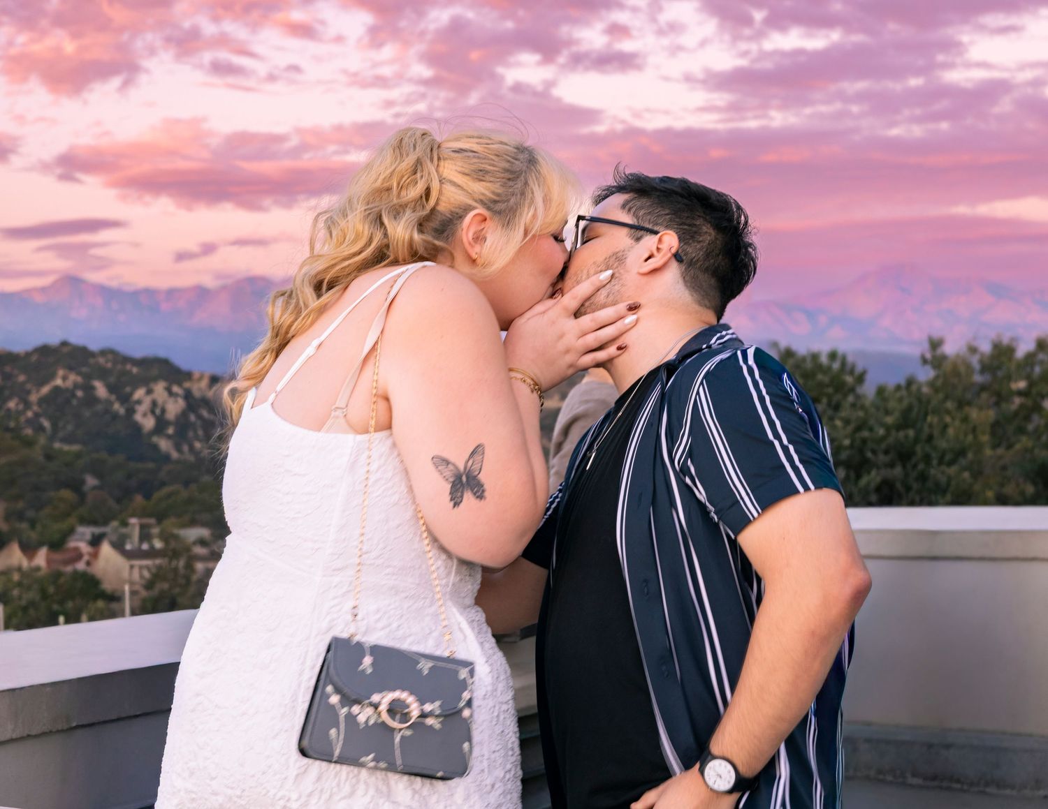 A romantic couple embraces during sunset with mountains and pink sky in the background.