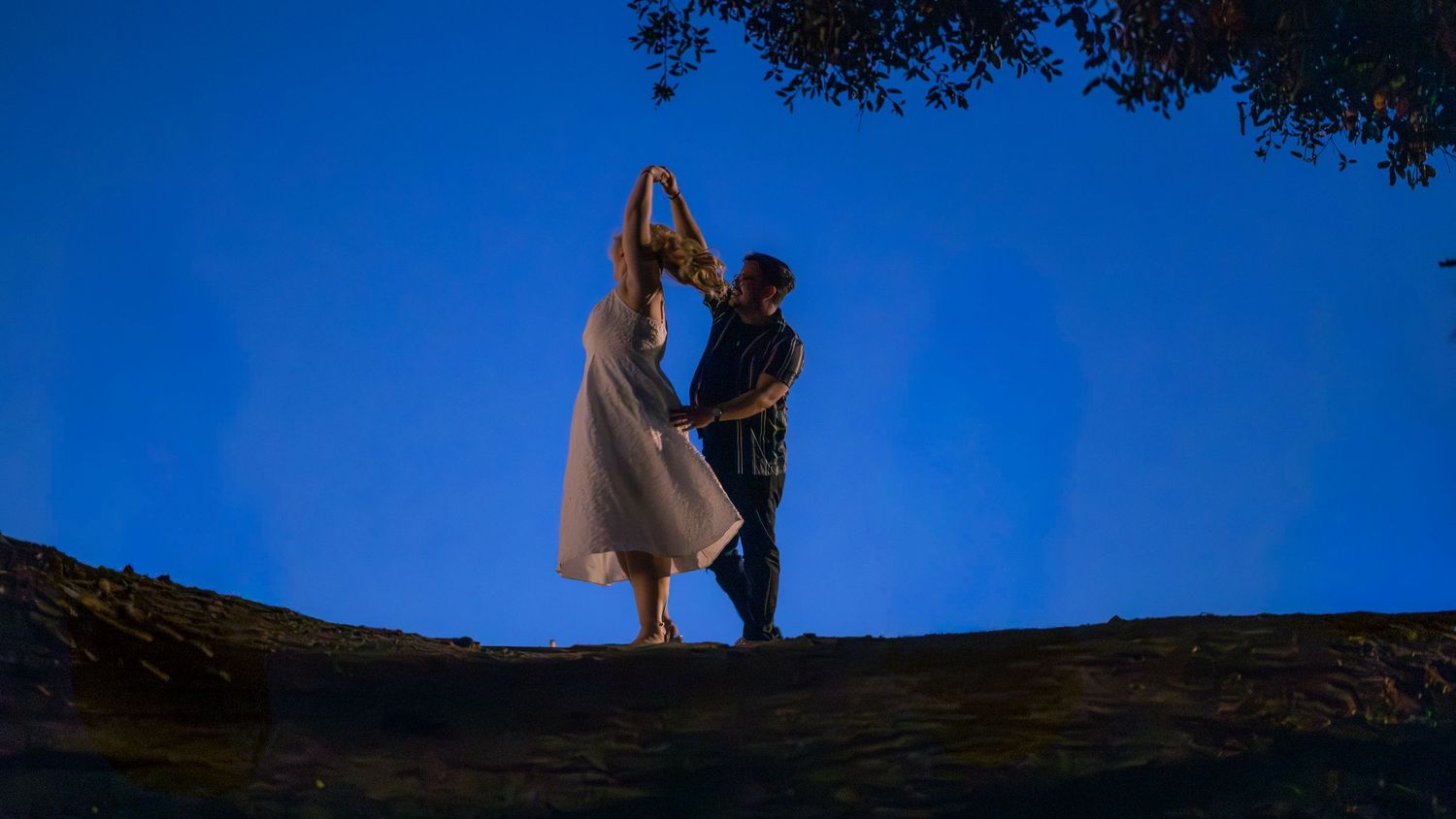 A silhouetted couple dances against a deep blue twilight sky beneath overhanging tree branches.
