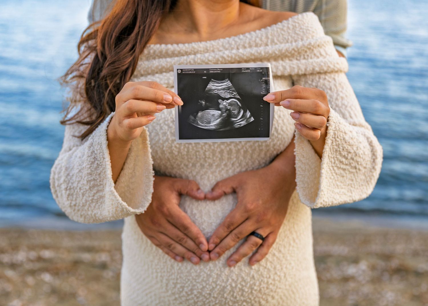 Pregnant woman in cream sweater holds ultrasound photo on beach during sunset pregnancy announcement photoshoot.