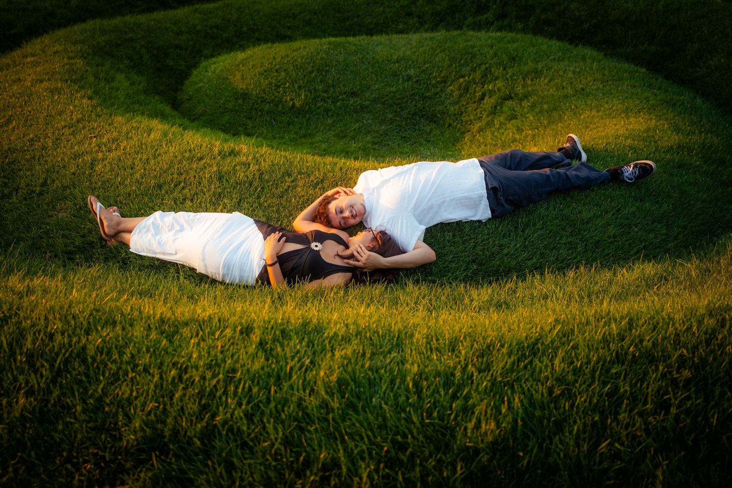 Couple lying on spiral grass mound, embracing romantically in lush green landscape.