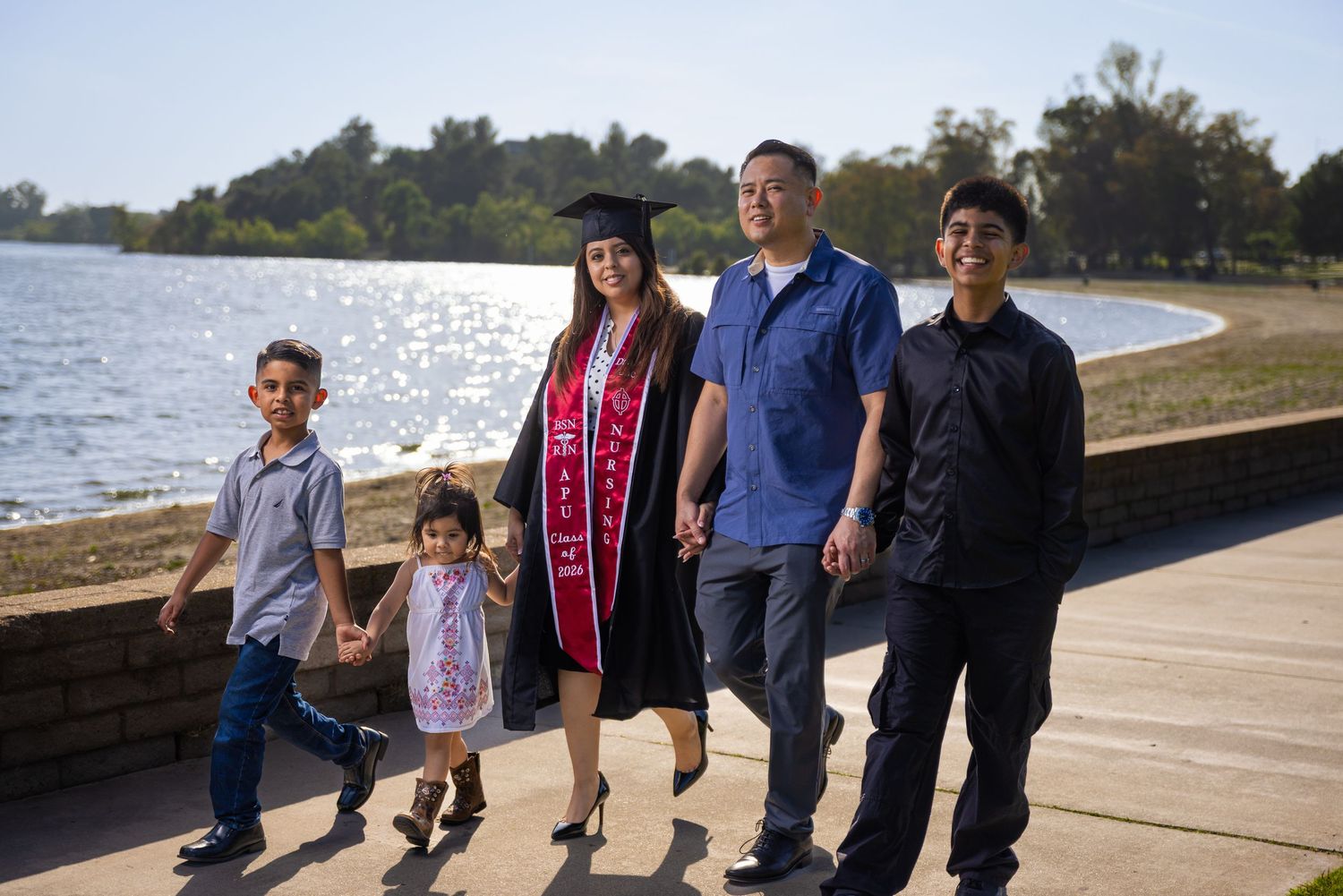 Family walking outdoors near a lake, celebrating a graduation with cap, gown, and red sash.