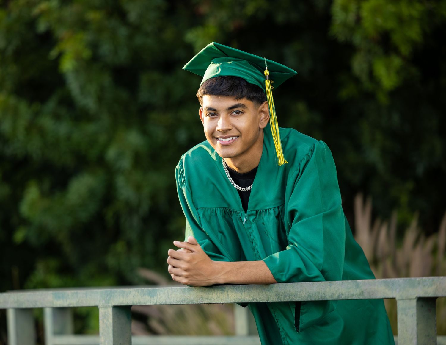 Smiling young man in green graduation cap and gown leaning on a railing outdoors with trees in background.
