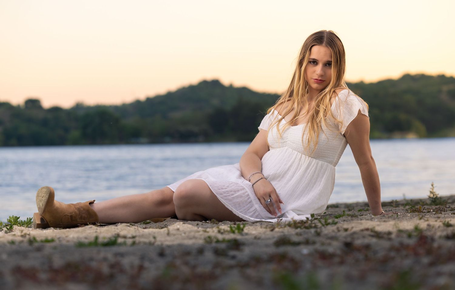 Pregnant woman in white dress and boots sitting on sandy beach at sunset near a river.