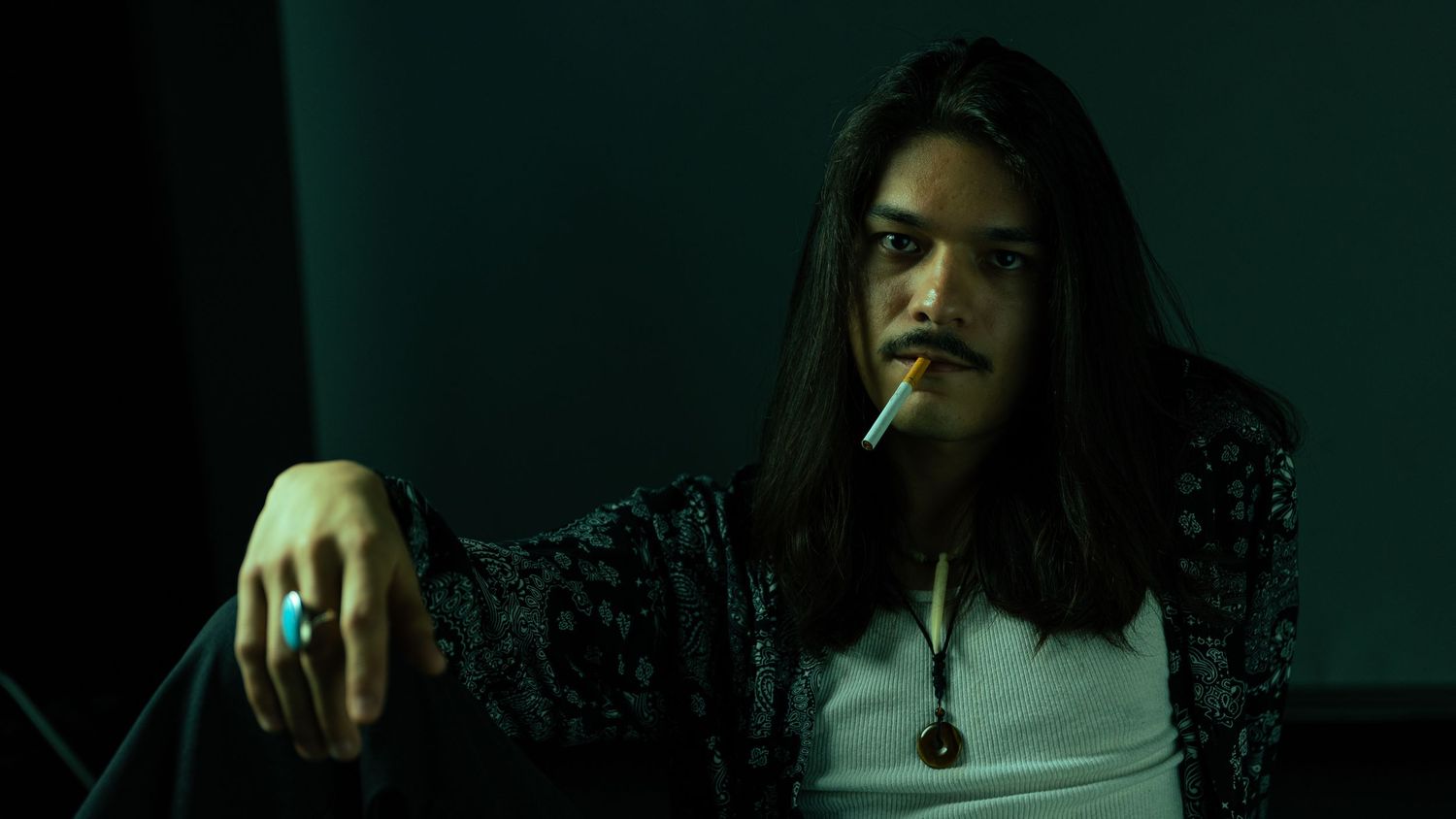 A person with long dark hair holds a cigarette in a dimly lit room with a moody green atmosphere.