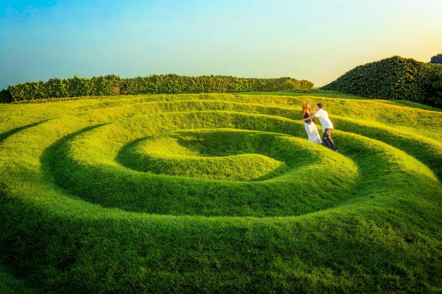 A couple walks through a stunning spiral-shaped green grass maze under a clear blue sky.