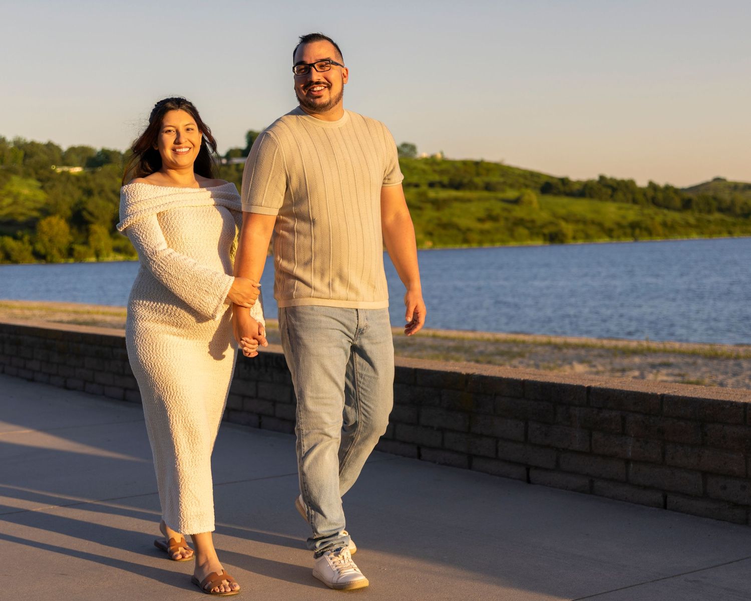 A couple in white outfits walking together along a waterfront path at sunset.