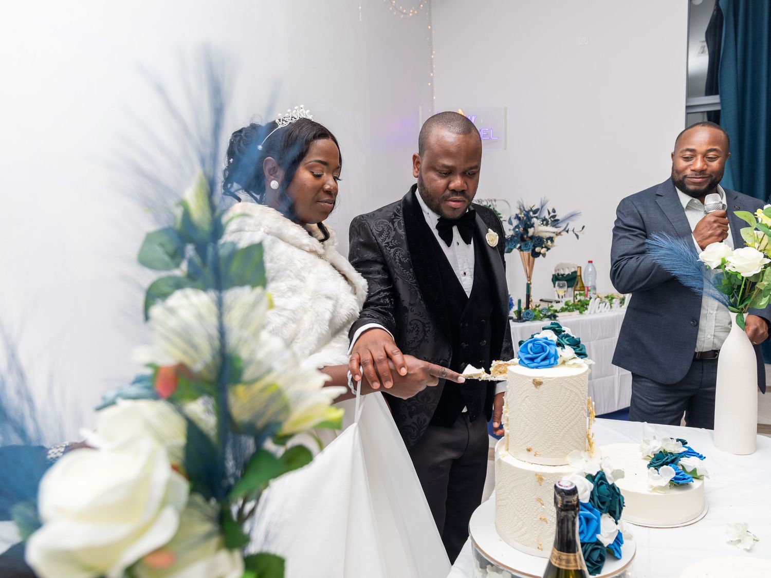 Bride and groom cutting white wedding cake decorated with blue flowers at reception celebration.