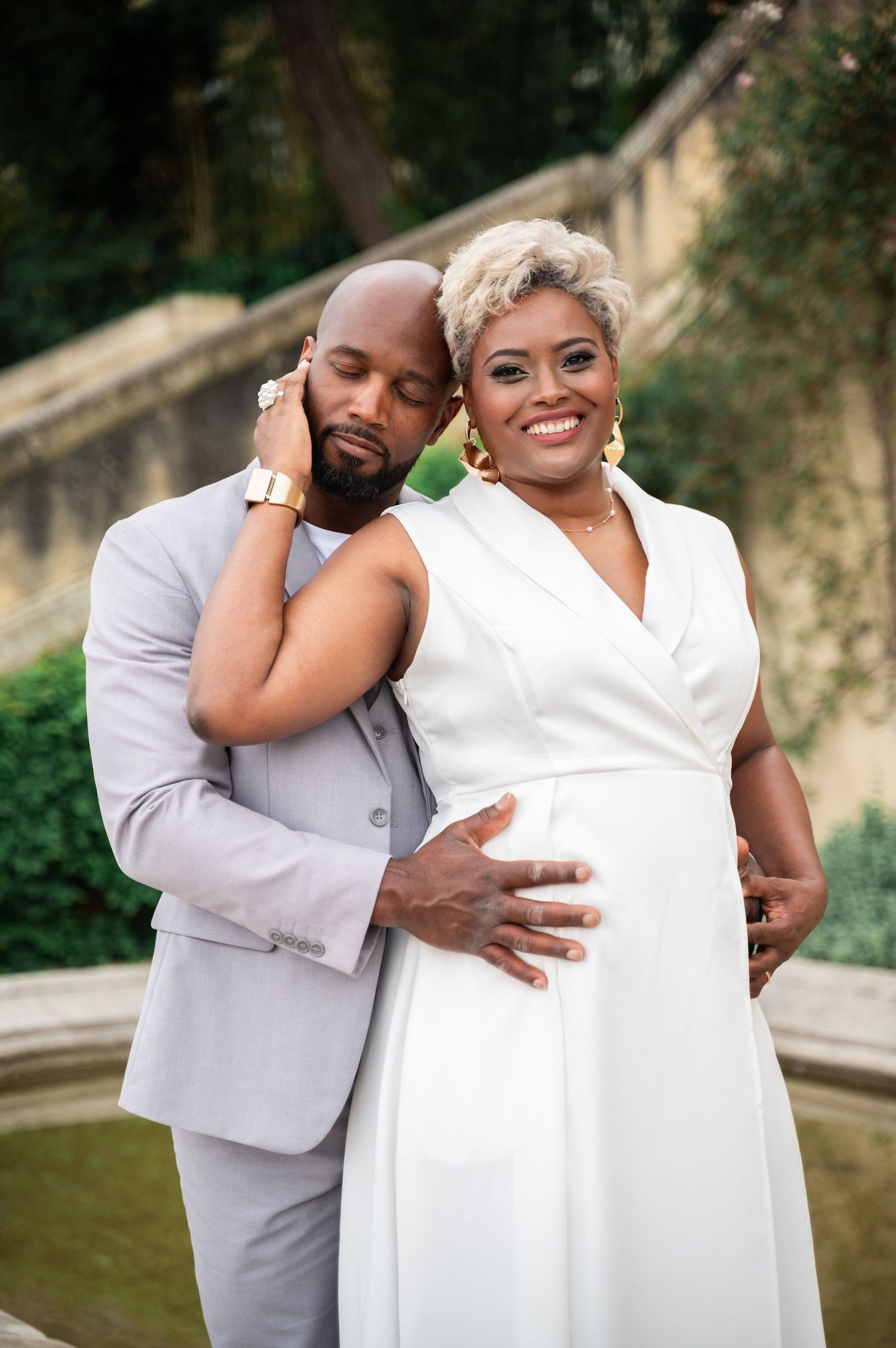 Black couple embracing near fountain, man in gray suit resting head on woman in white dress.