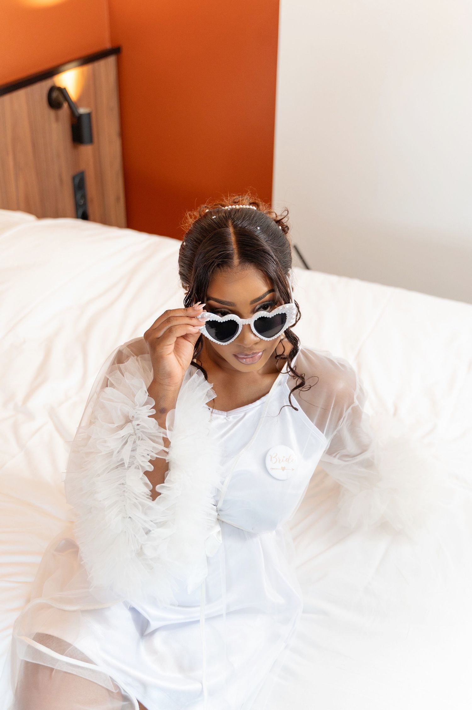 Black bride in white robe wearing heart-shaped sunglasses sitting on hotel bed.