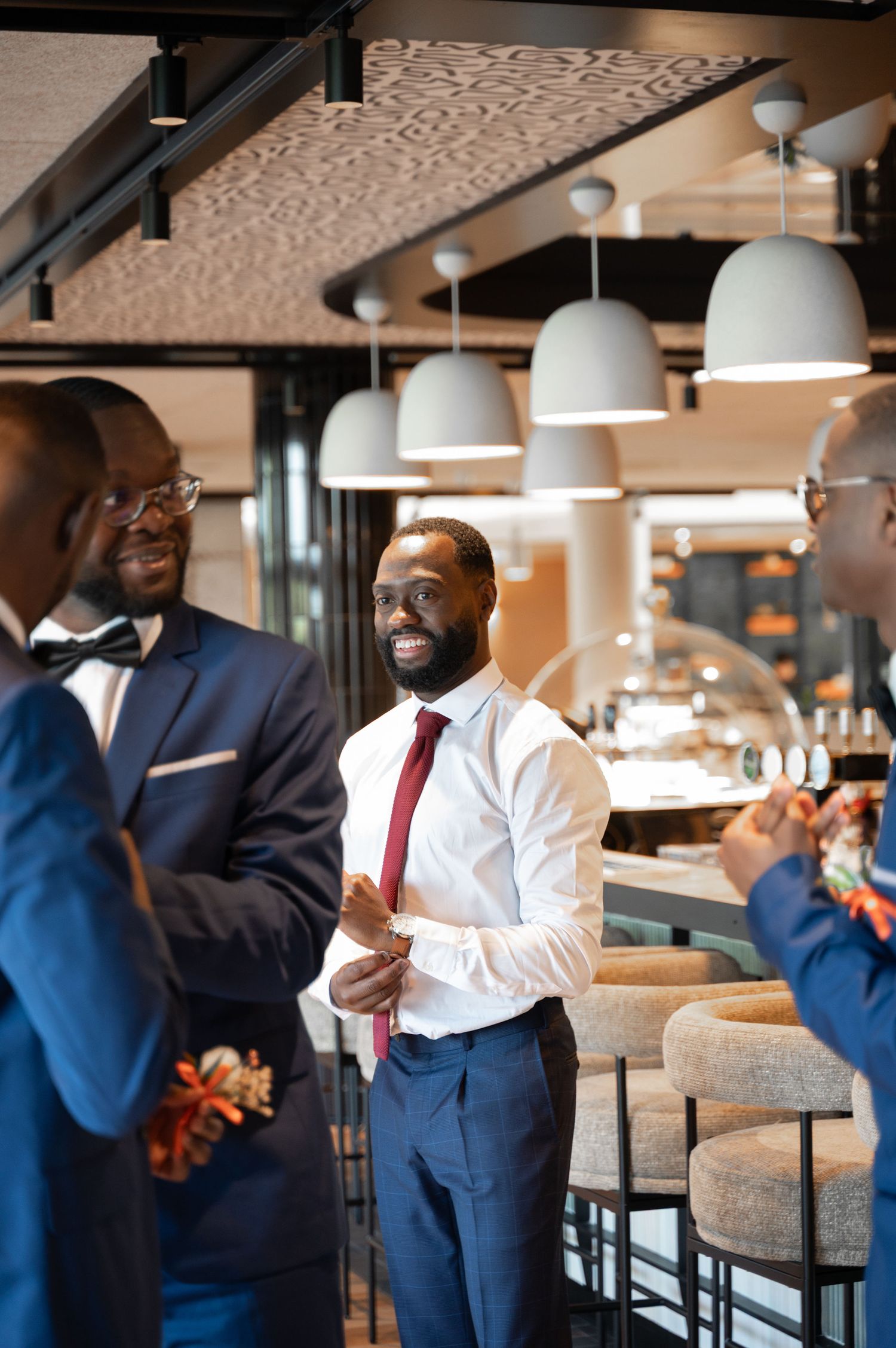 Handsome Black groom in white shirt and red tie smiles joyfully among groomsmen in blue suits.