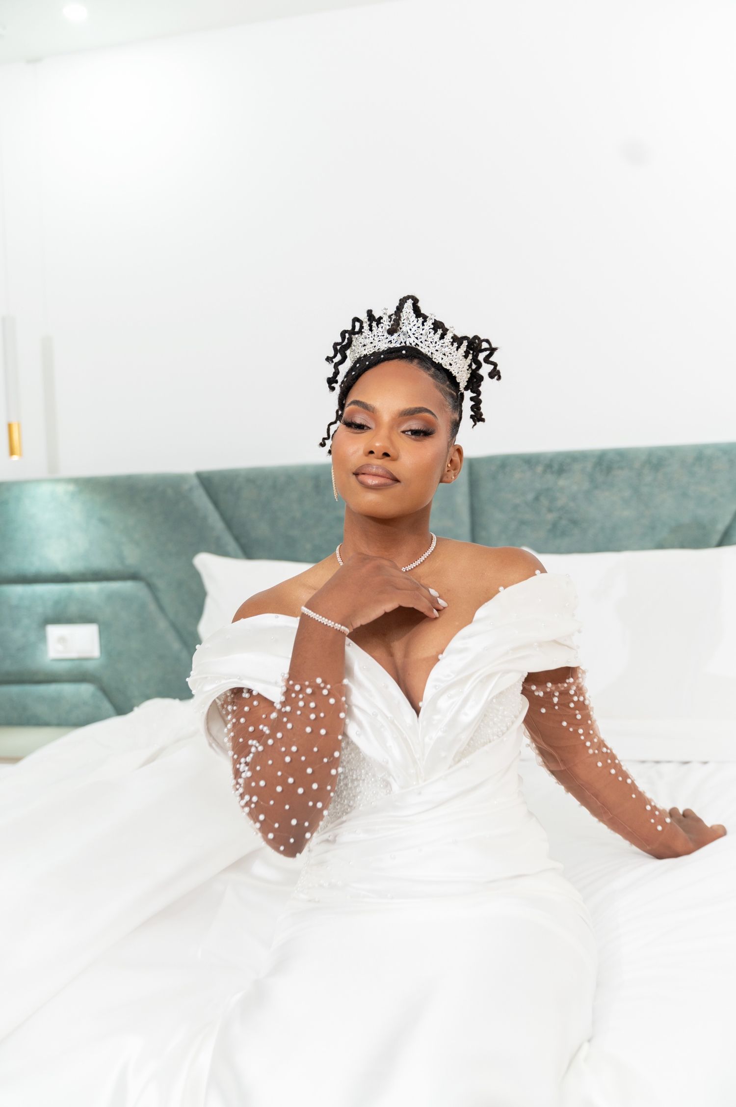 Elegant Black bride in pearl wedding gown and crystal tiara poses on white bed.