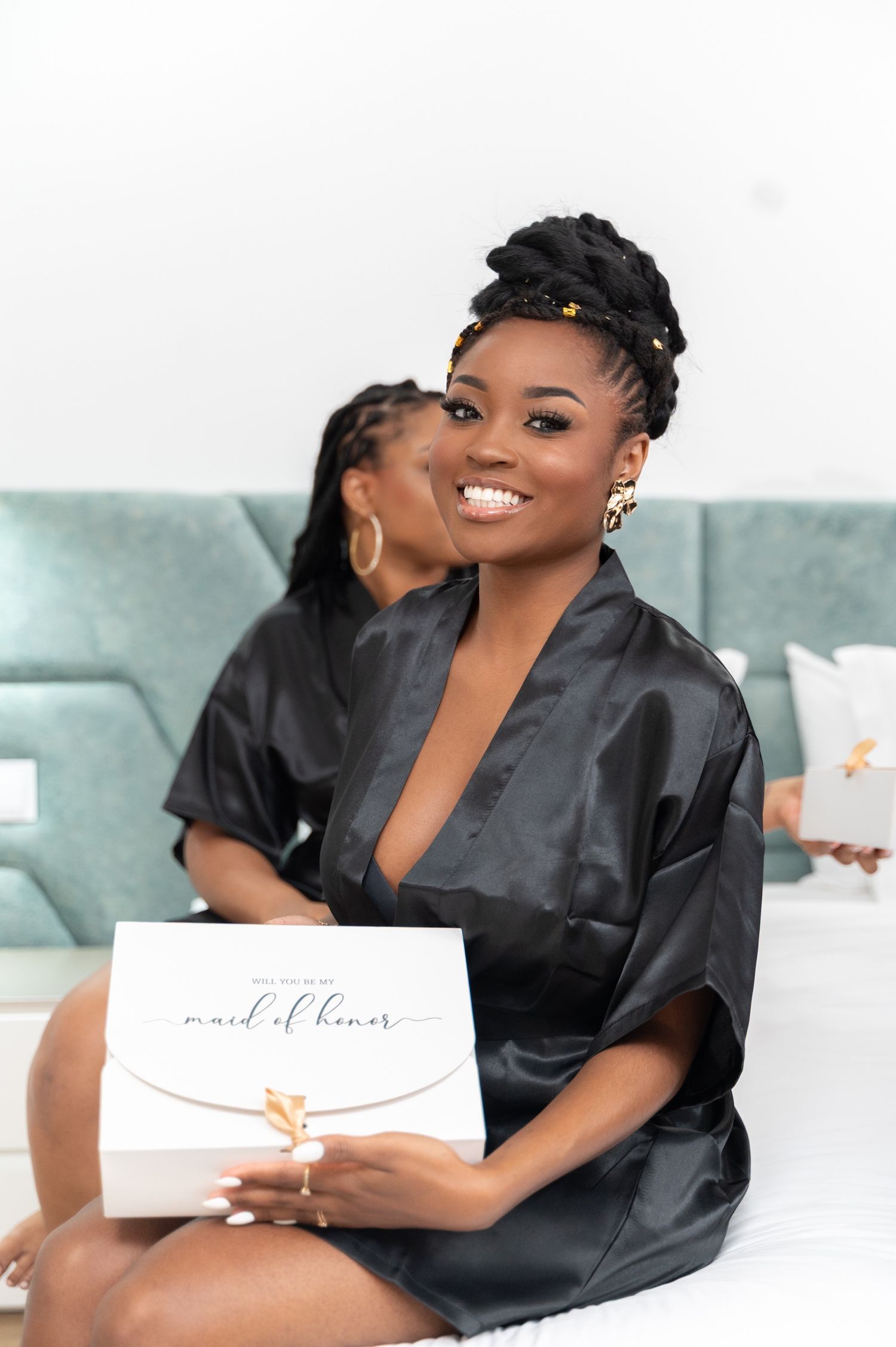 Smiling bridesmaid in black satin robe holding a maid of honor gift box while sitting on a bed.
