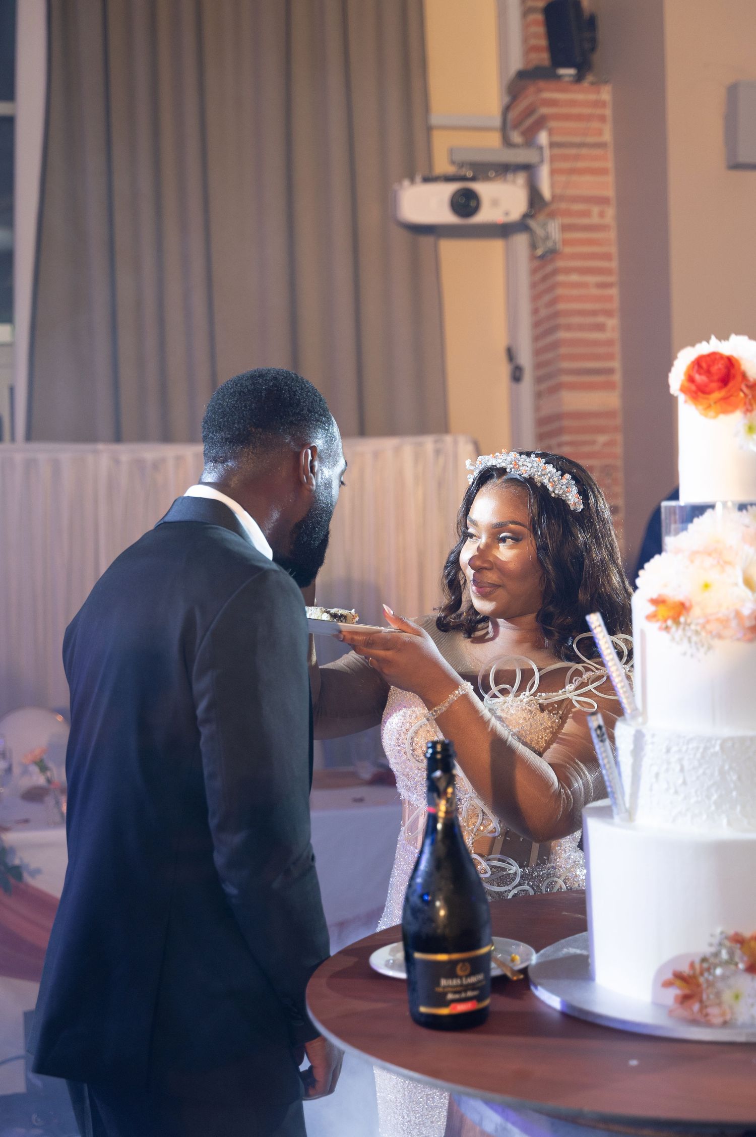 Bride in beaded gown feeds groom cake while champagne bottle sits on barrel table at reception.