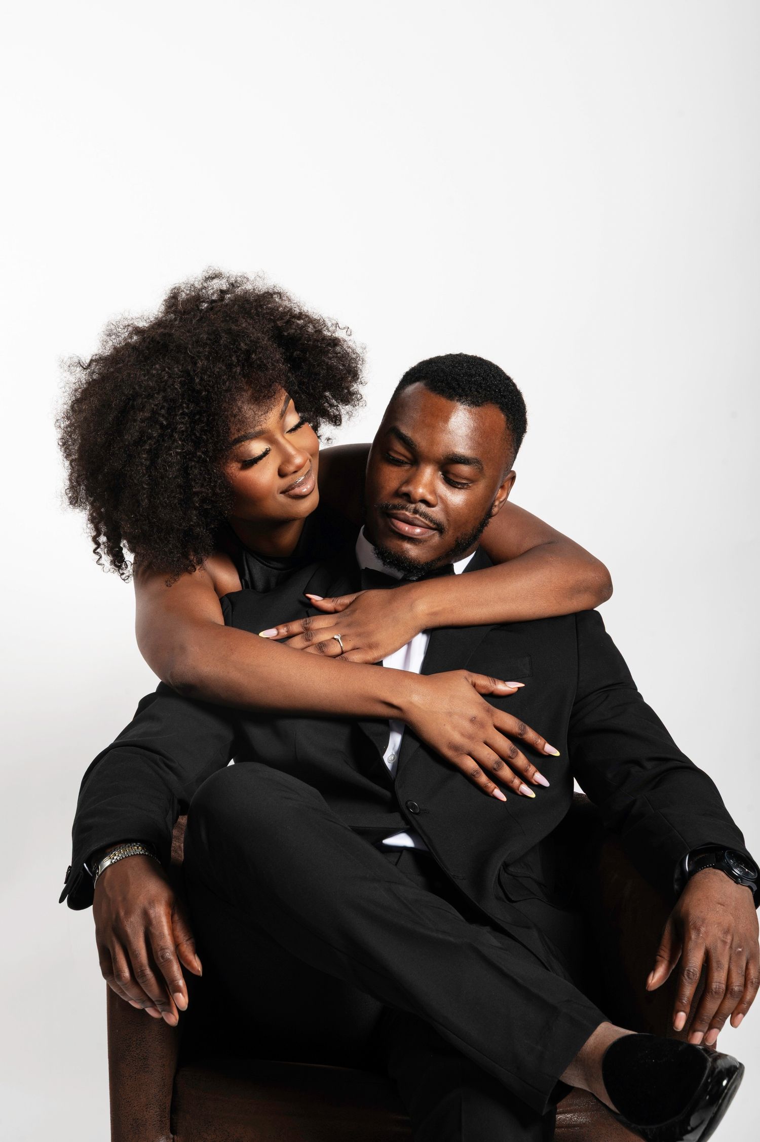 A woman in a black dress embracing a seated man in a black suit against a white background.