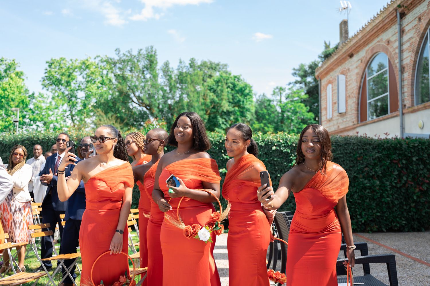 Group of bridesmaids in matching orange one-shoulder gowns smiling at outdoor wedding ceremony.