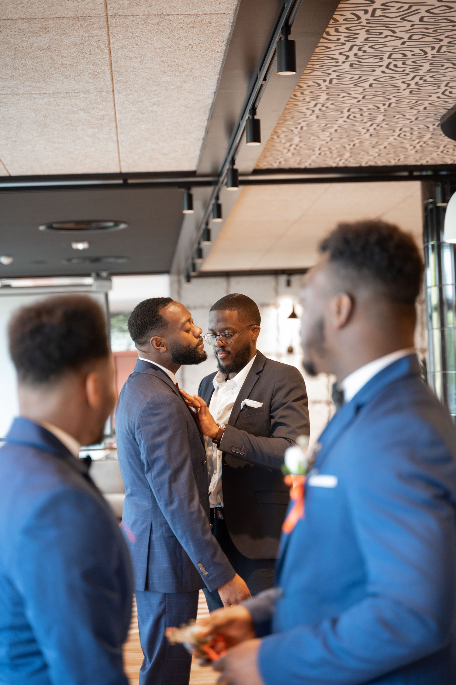 Group of Black men in blue suits gathered indoors as one adjusts another's tie before a wedding ceremony.