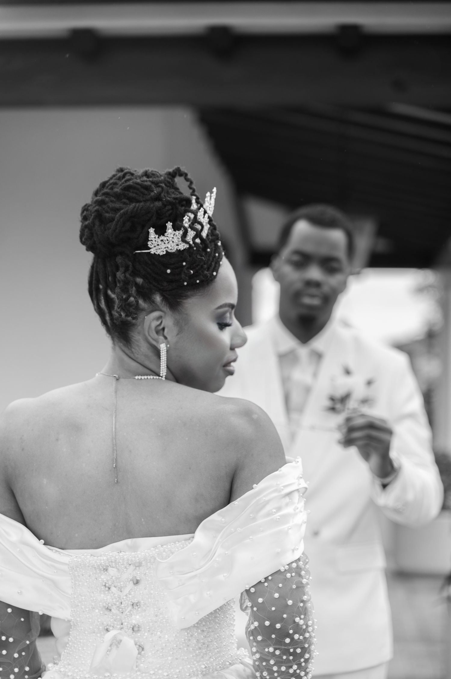 Black and white photo of a bride in a beaded gown and crown tiara with groom in white suit behind her.