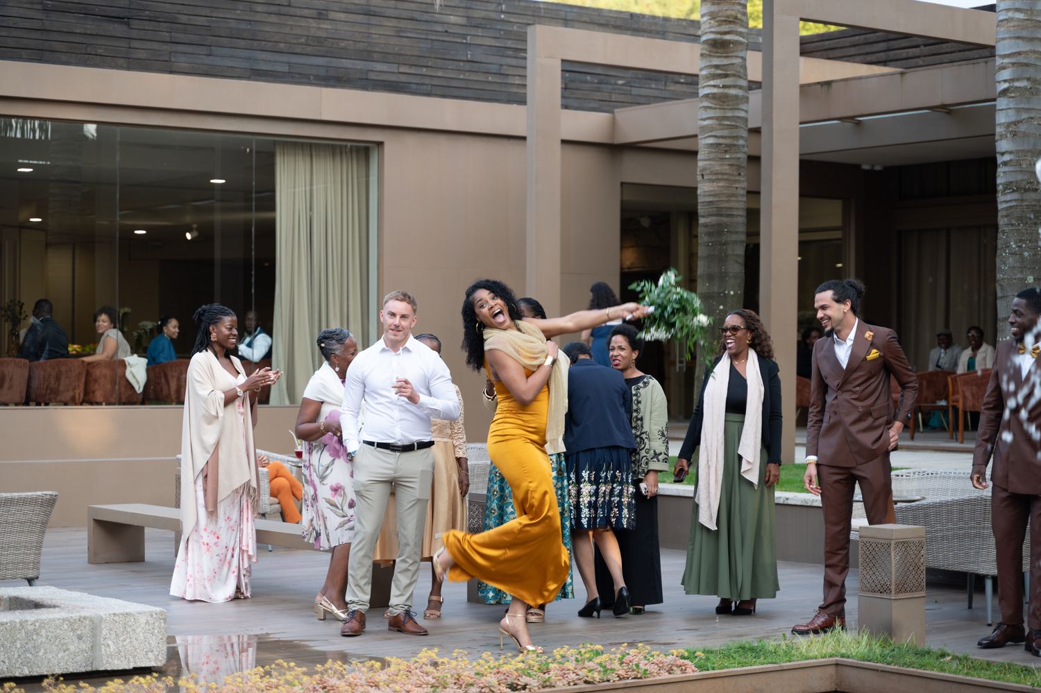 Wedding guests celebrate outdoors as woman in yellow dress dances joyfully on decorative patio at venue.