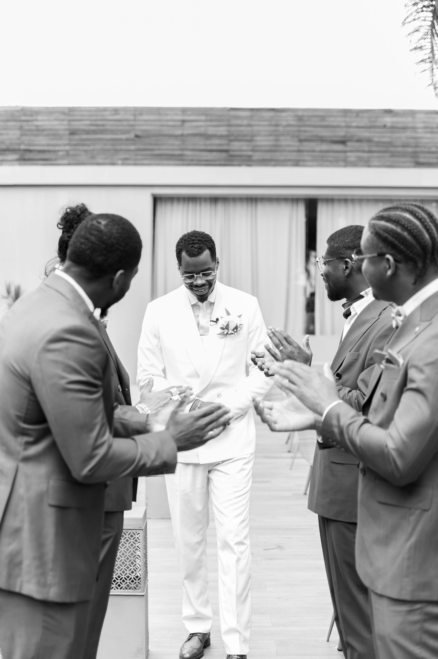 Groomsmen in brown suits applaud the groom in a white suit during a wedding ceremony in black and white.