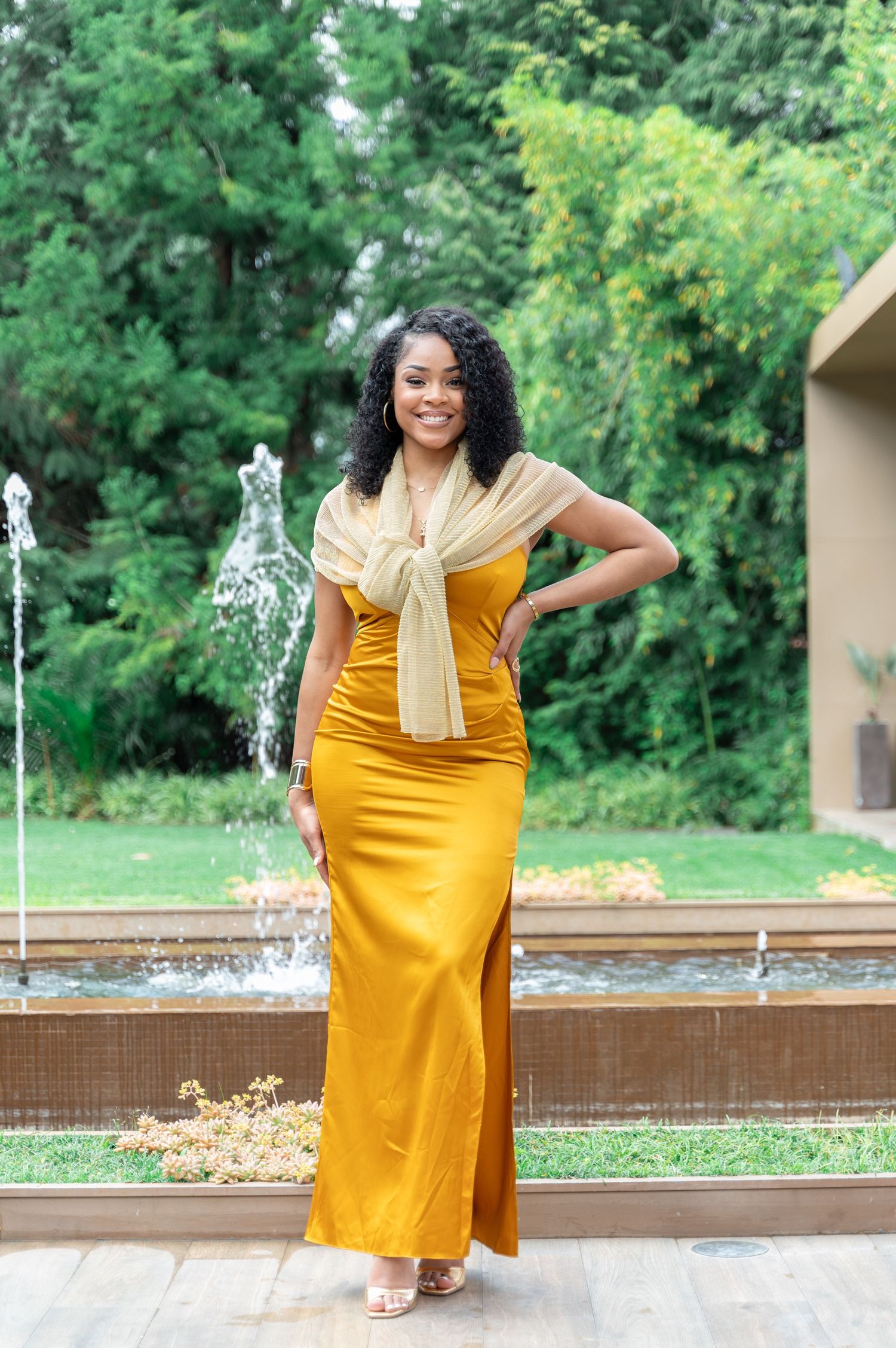 A woman in a golden yellow maxi dress with a matching scarf poses elegantly in front of a garden fountain.