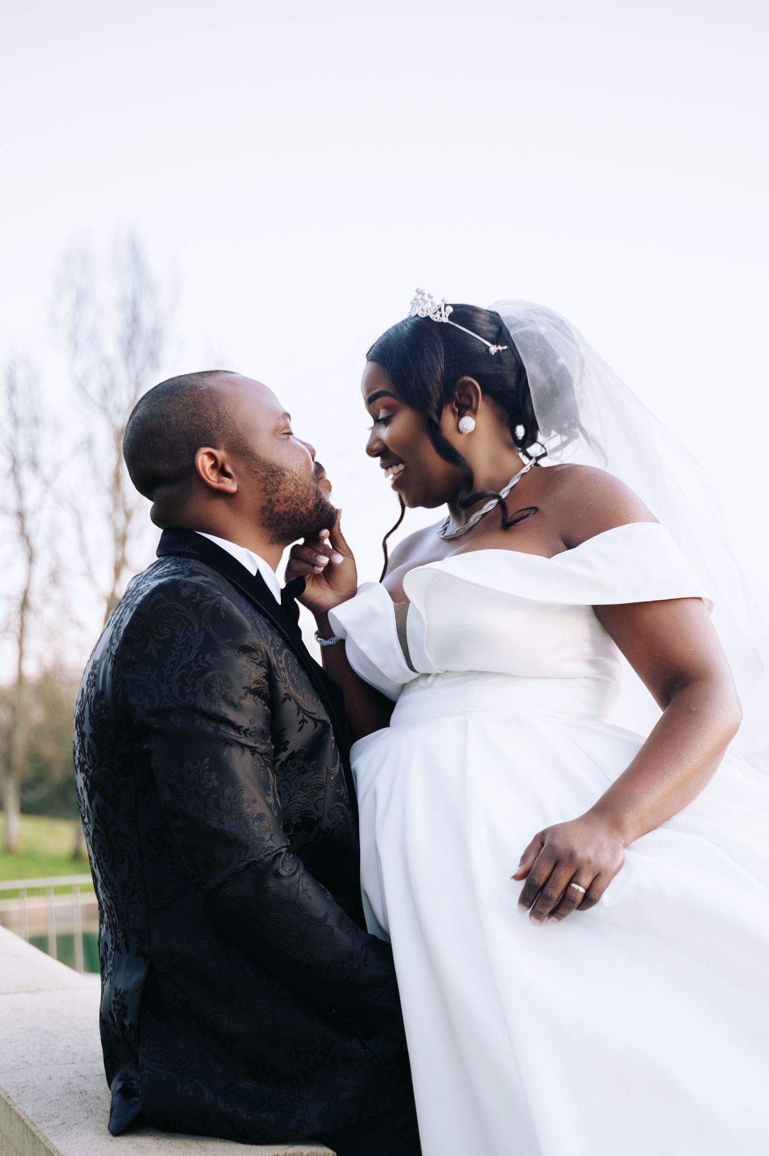 A Black bride and groom share a romantic moment, with the bride in a white off-shoulder gown and veil, touching the grooms face.