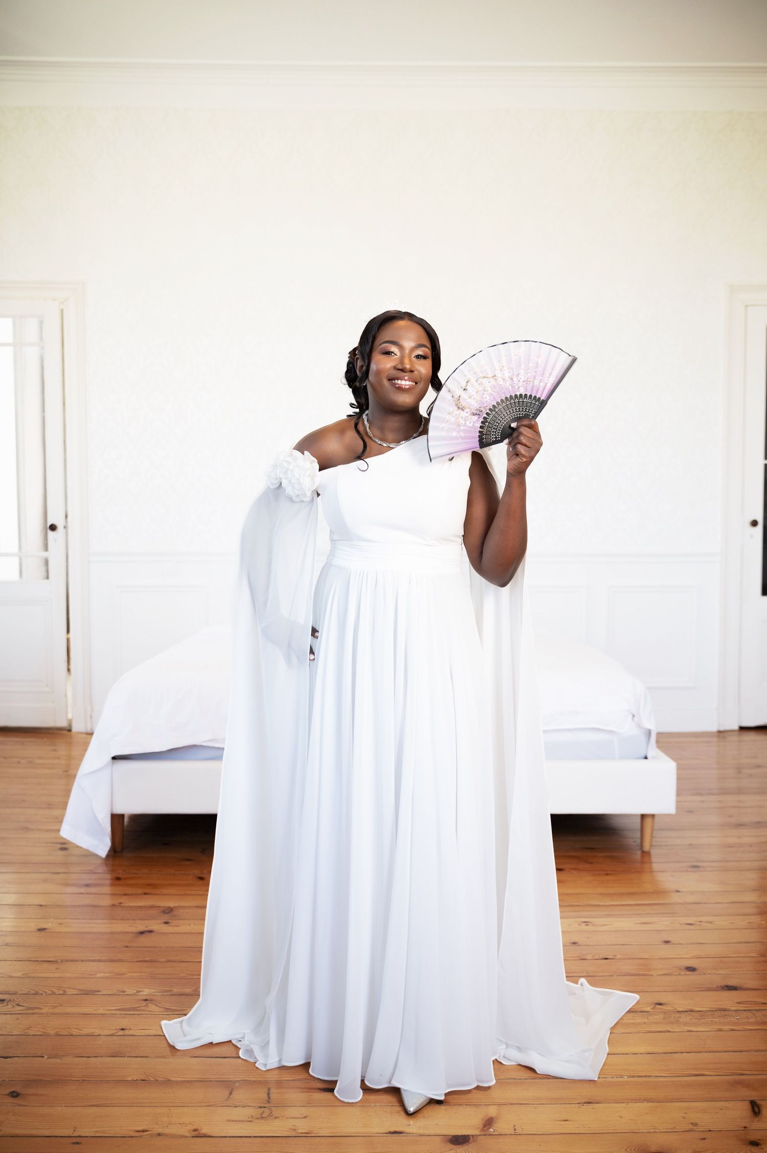 Bride in white off-shoulder gown holding decorative fan in bright bedroom setting.