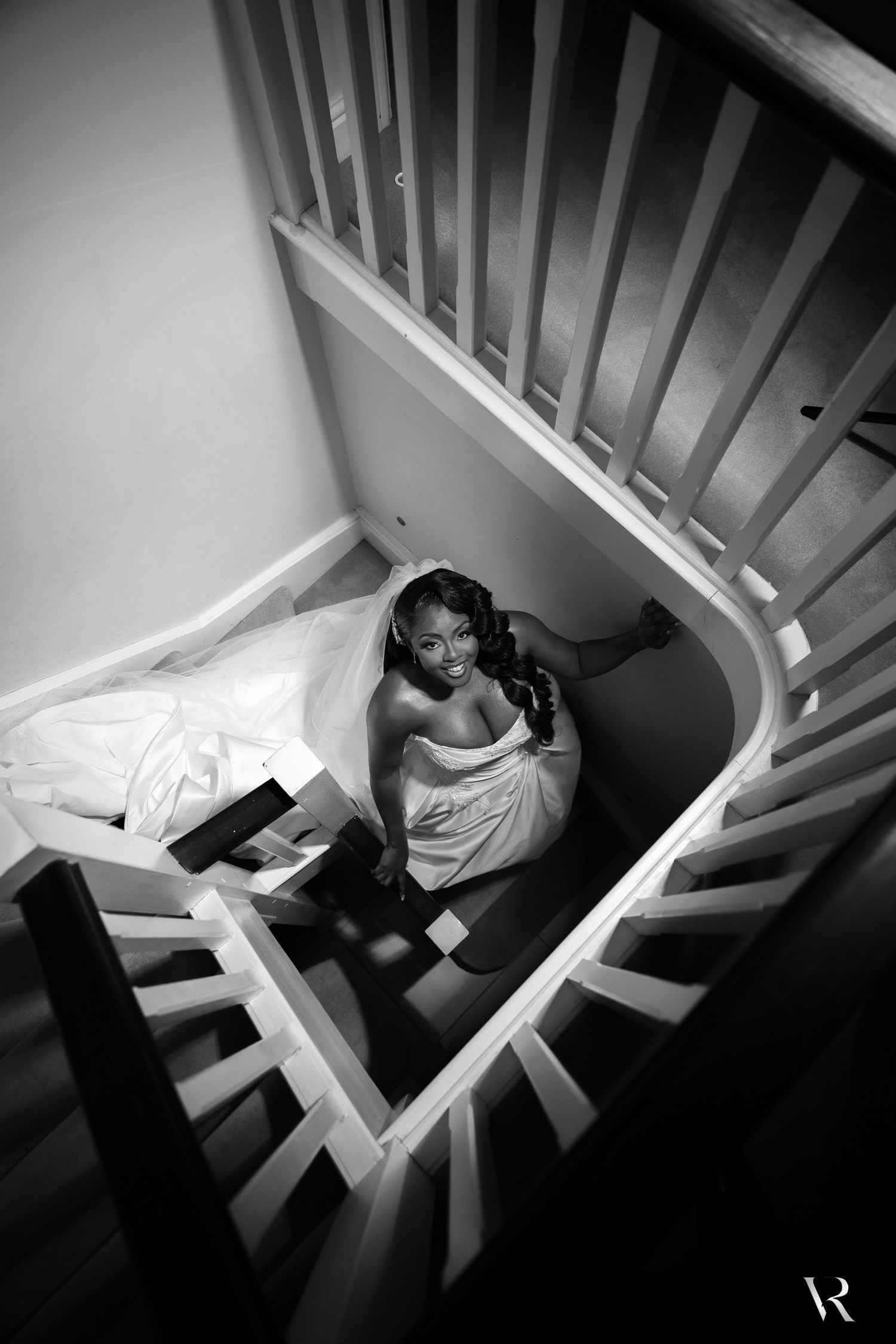 Black and white aerial view of a bride in a white gown sitting on a spiral staircase, looking up at the camera.