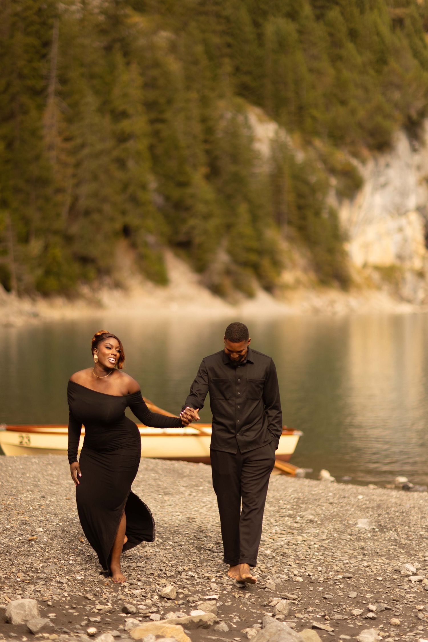 A couple dressed in black walks hand-in-hand along a rocky lakeside shore with a wooden boat and pine trees behind them.