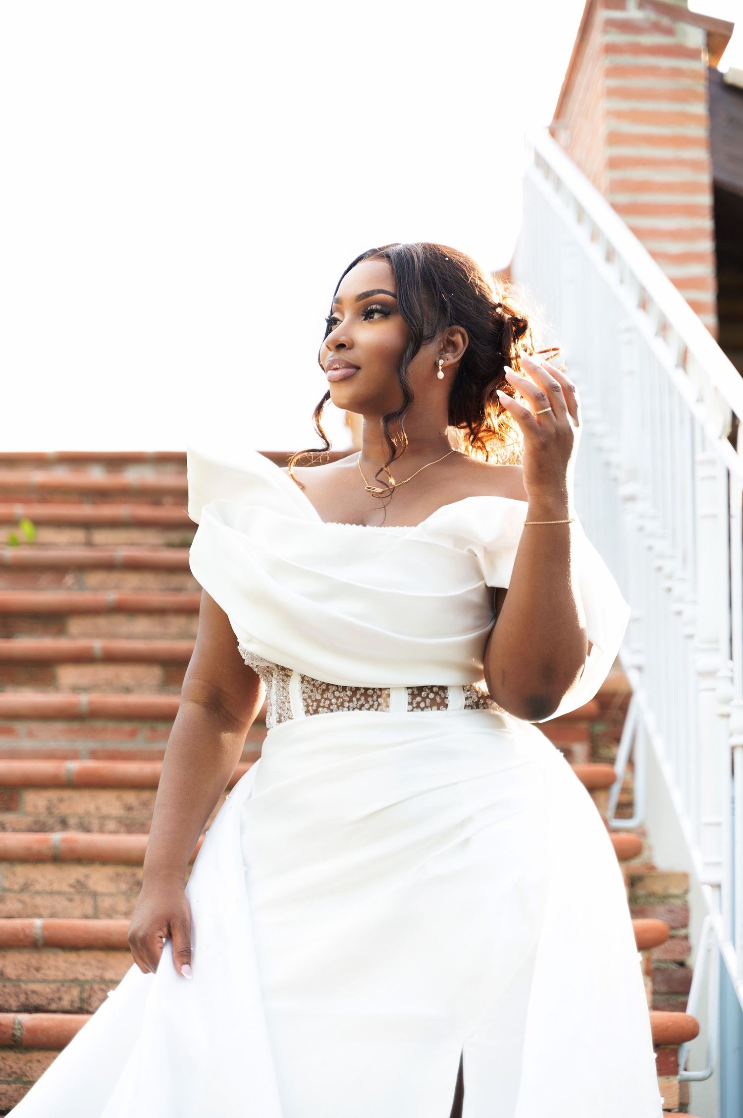 Stunning bride in white off-shoulder gown with gold waist detail posing gracefully on brick stairs.