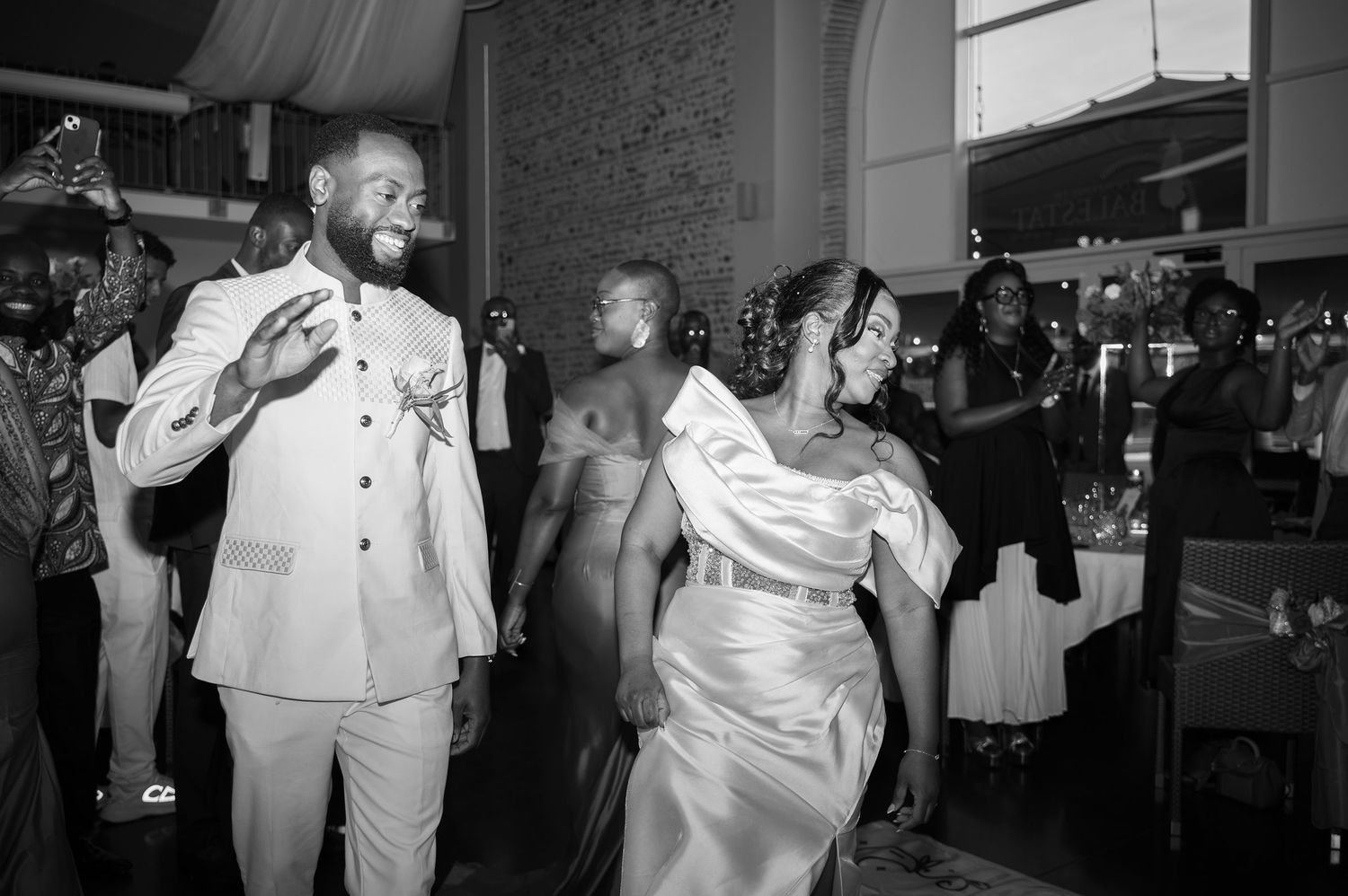 Black and white photo of smiling groom in white suit and bride entering wedding reception.