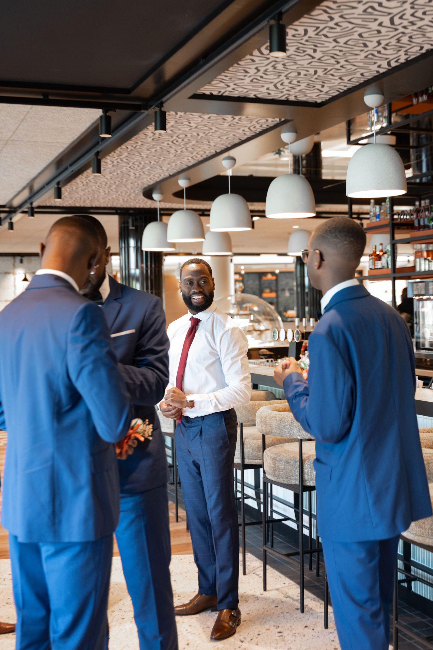 Three well-dressed Black men in blue suits chat and laugh together inside a modern restaurant bar venue.