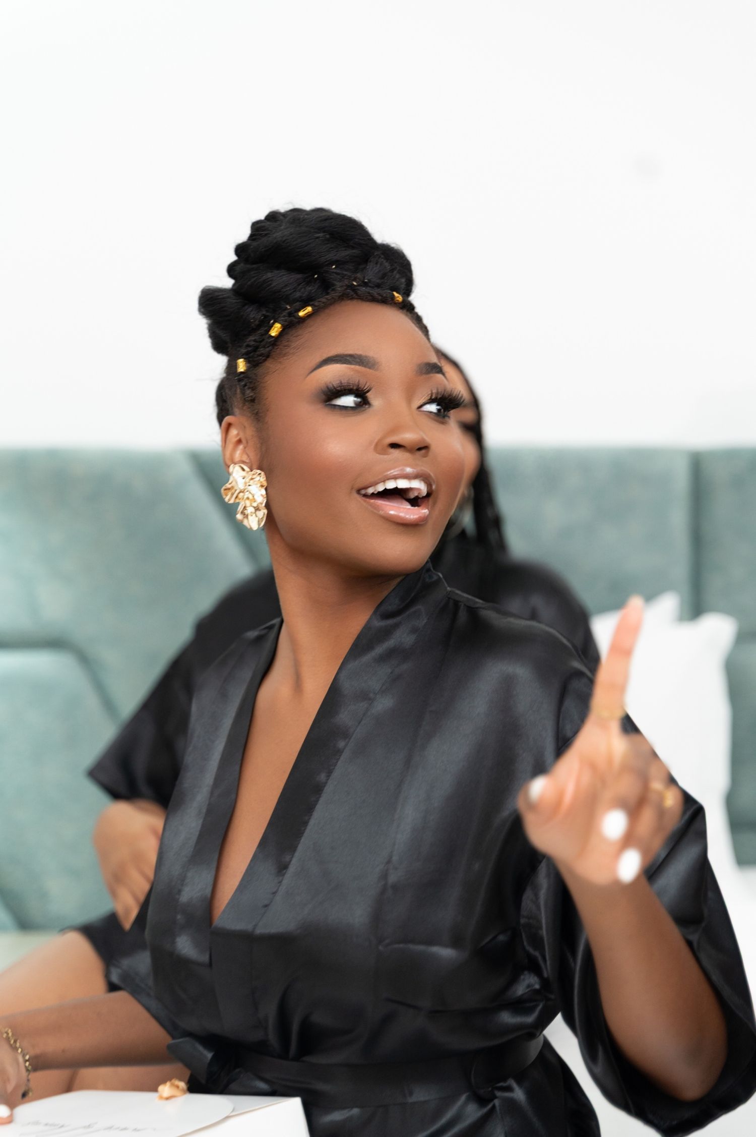 Joyful woman in black satin robe with gold earrings gesturing while sitting on a bed.