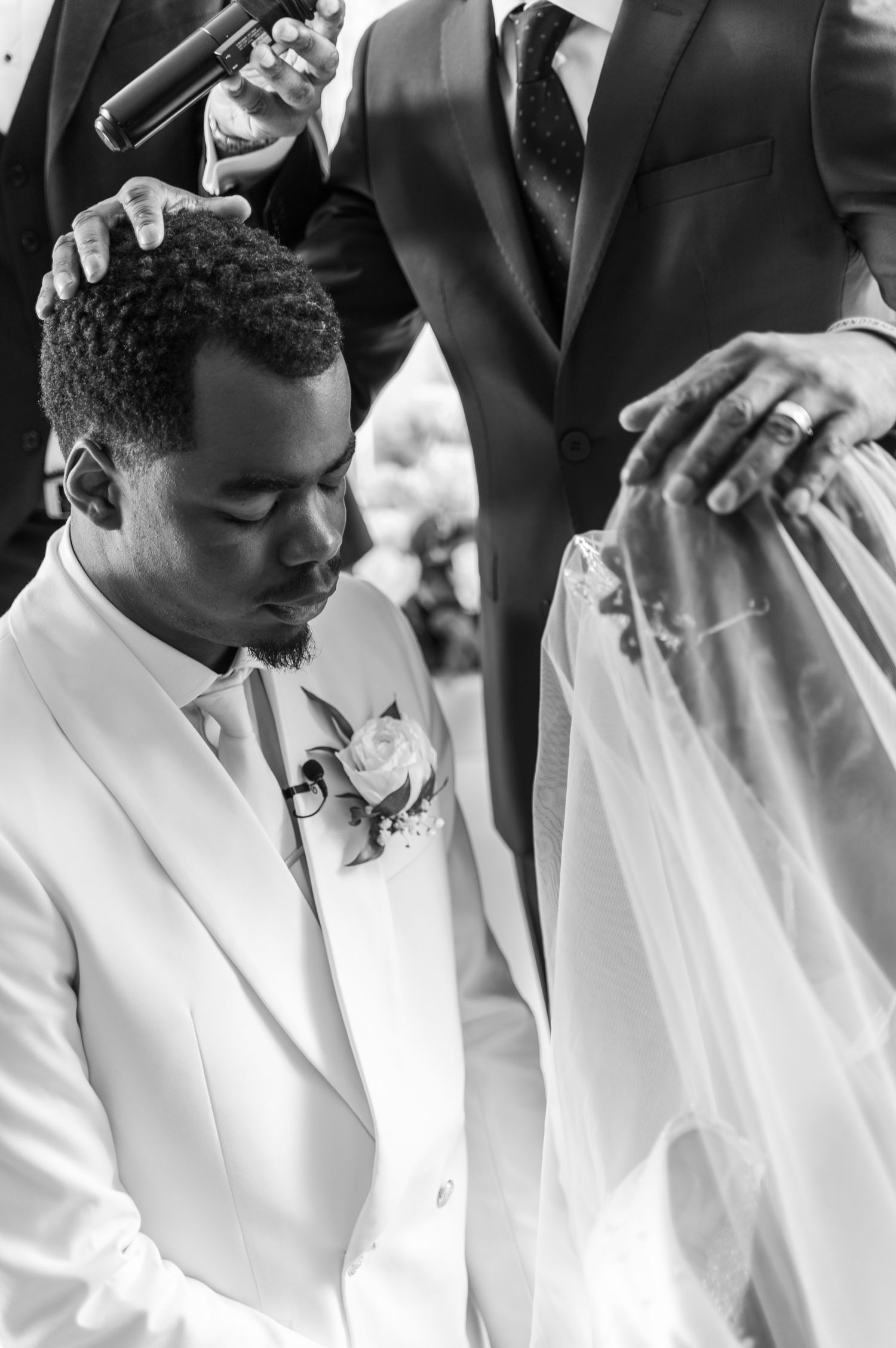 Black and white photo of groom bowing head in prayer during wedding ceremony blessing.