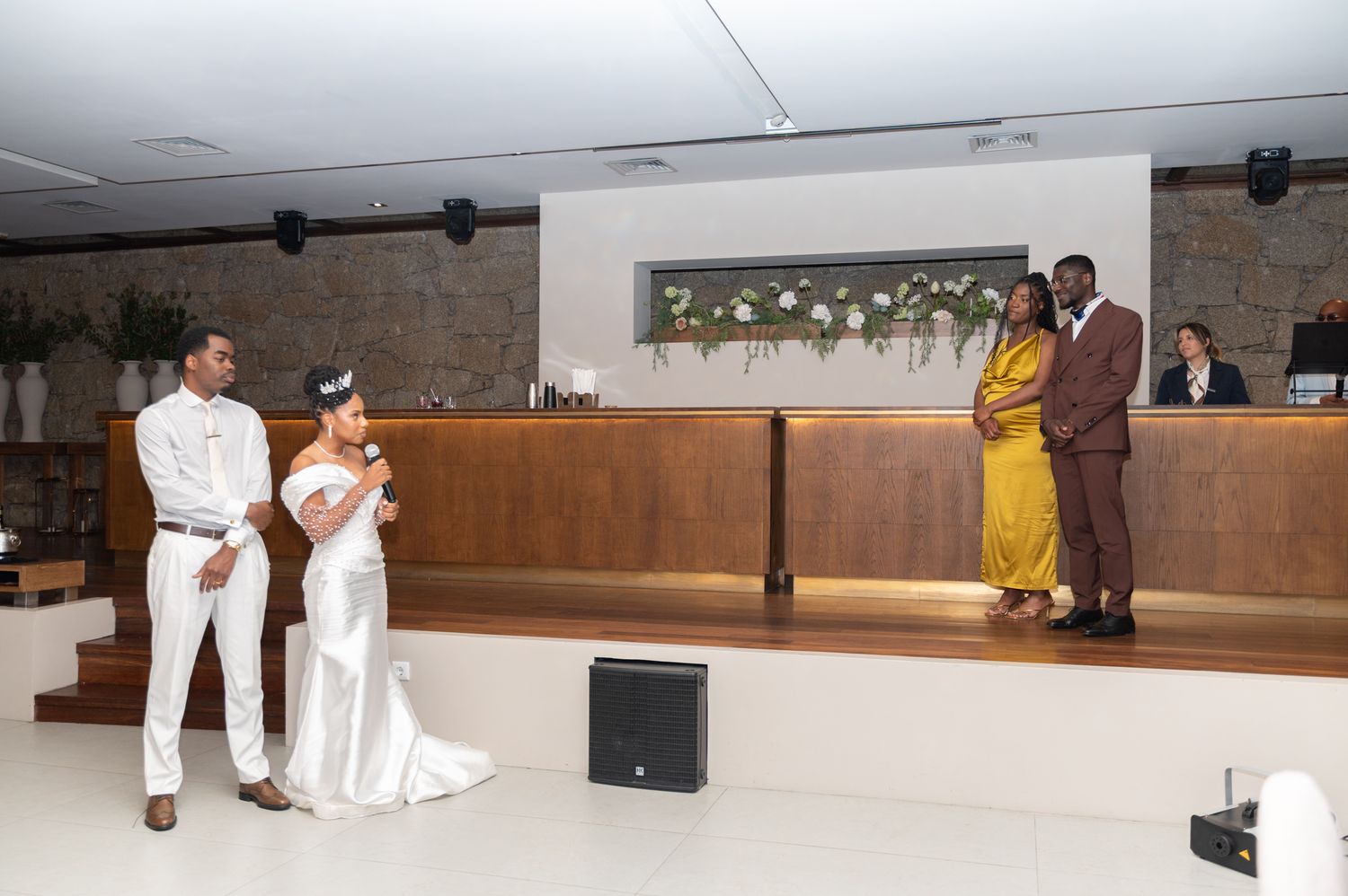 Wedding couple in white attire stands at altar while guests in gold and brown outfits look on indoors.