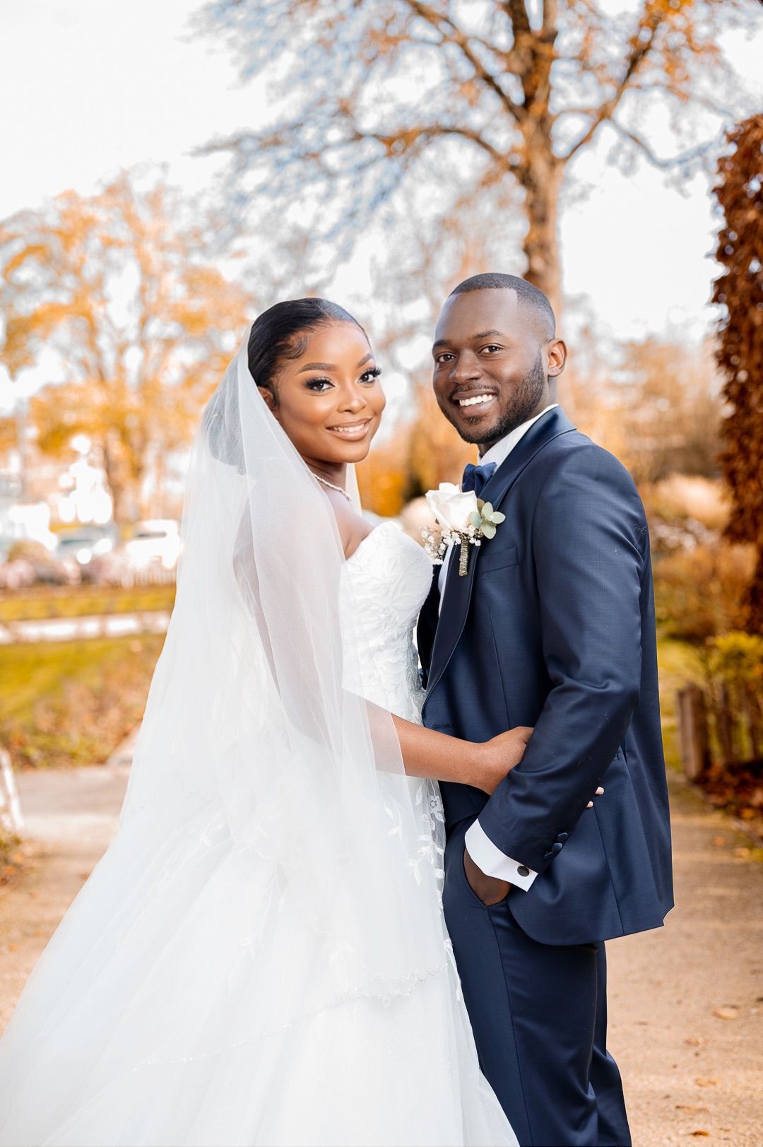Happy Black bride and groom posing outdoors in autumn, she in white gown and veil, he in navy suit.