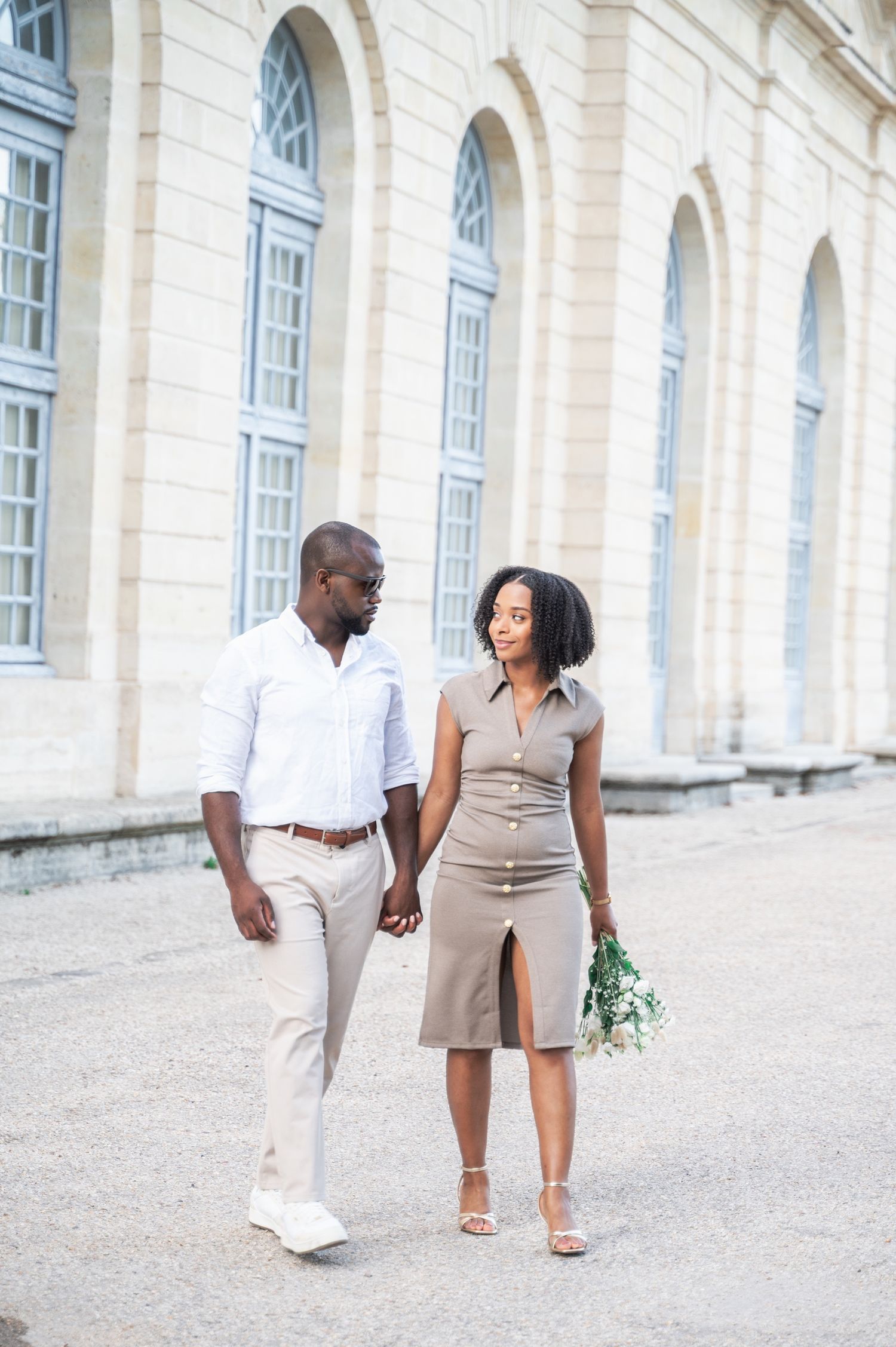An elegant couple walks hand in hand past a classical French building, the woman holding a white bouquet.