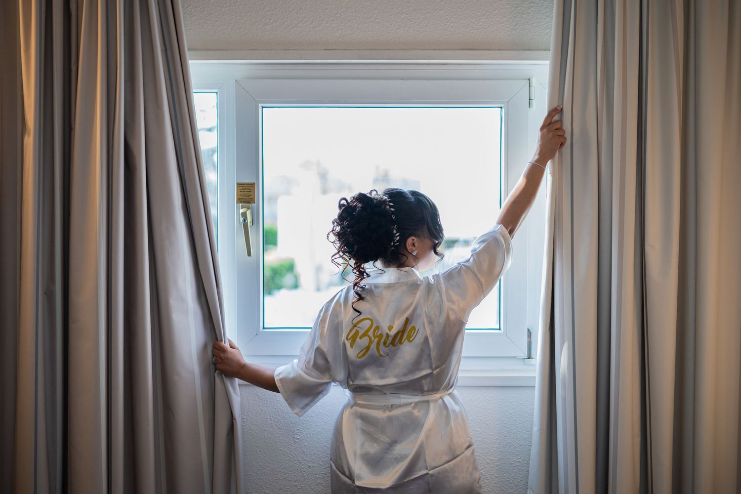 Bride in white satin robe with gold Bride text opens curtains facing a bright window.