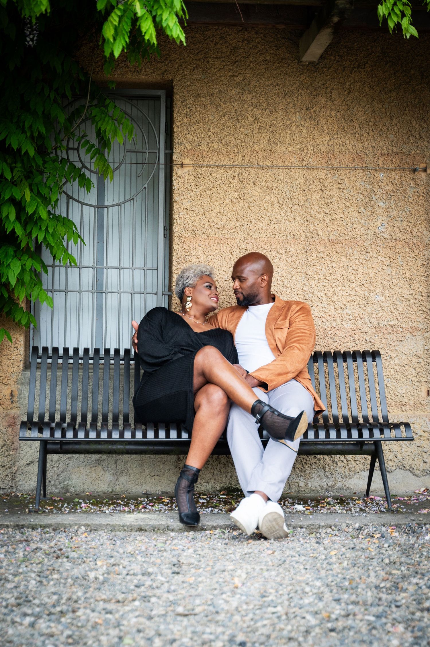 Black couple sitting close on bench, woman in black dress and man in tan jacket sharing intimate moment.