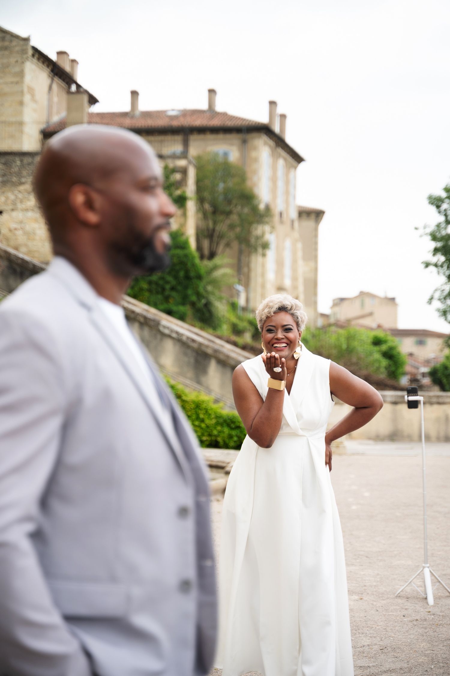 Bride in white jumpsuit smiling at groom in gray suit outdoors near a historic stone building.