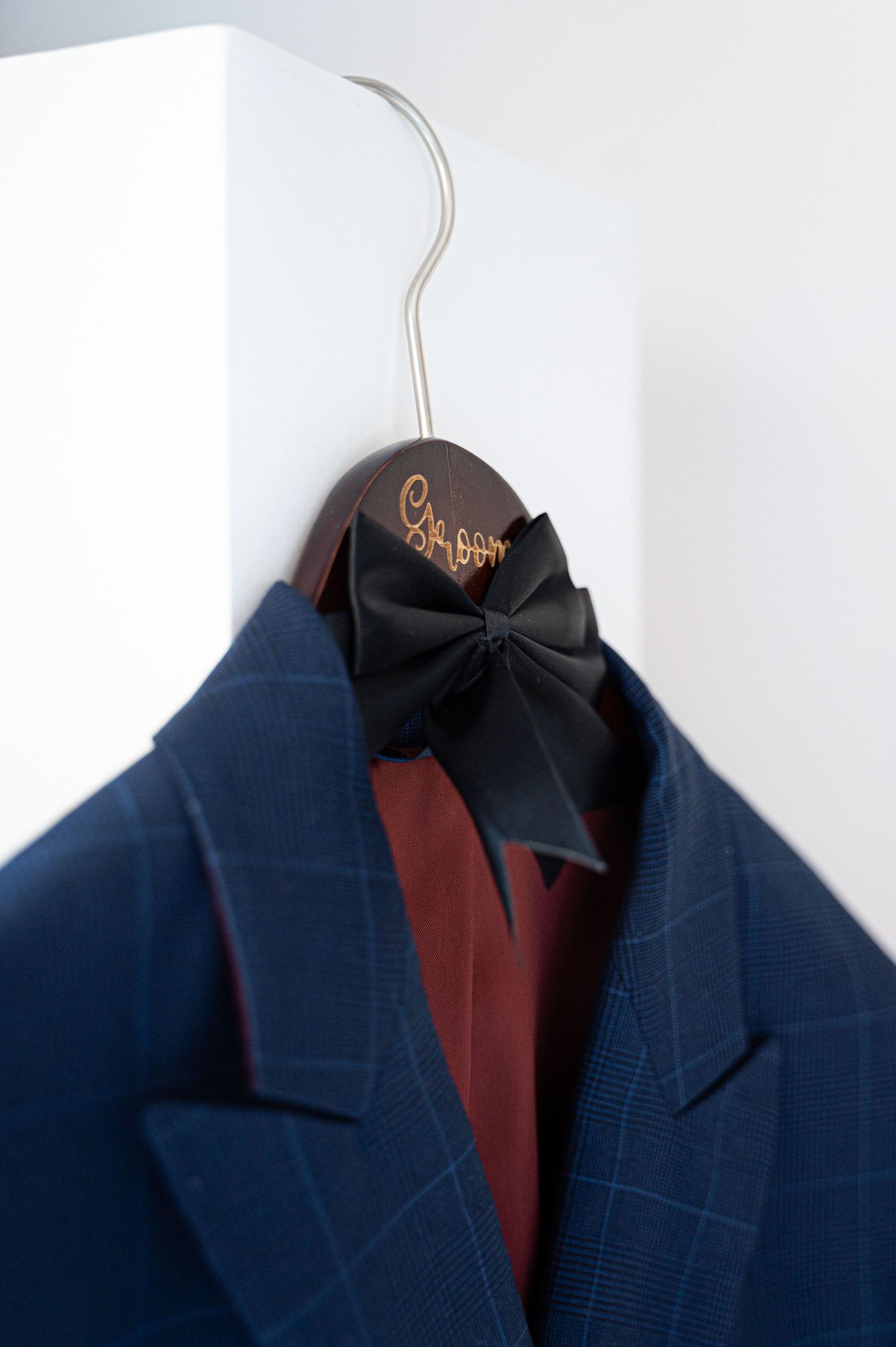 Navy blue plaid groom suit jacket on wooden hanger with black bow tie and burgundy shirt.