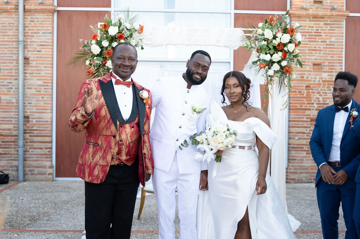 Bride and groom in white posing with smiling guest in red patterned jacket at outdoor wedding.