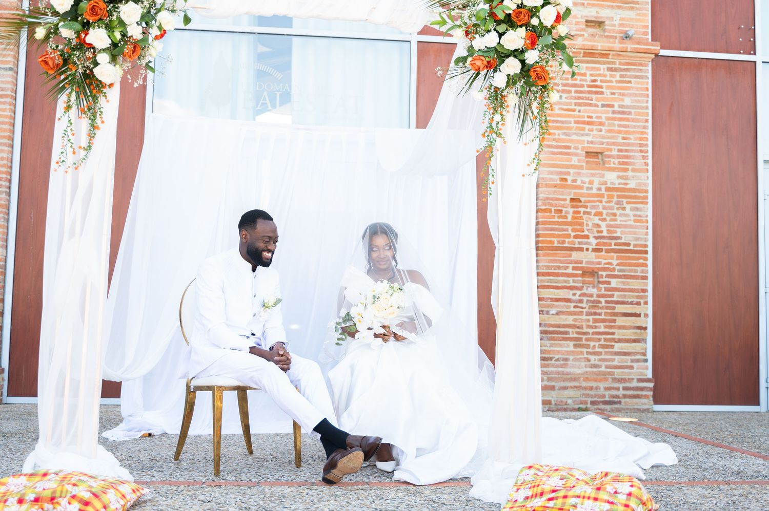 Bride and groom in white sit together under draped canopy with orange floral arrangements.