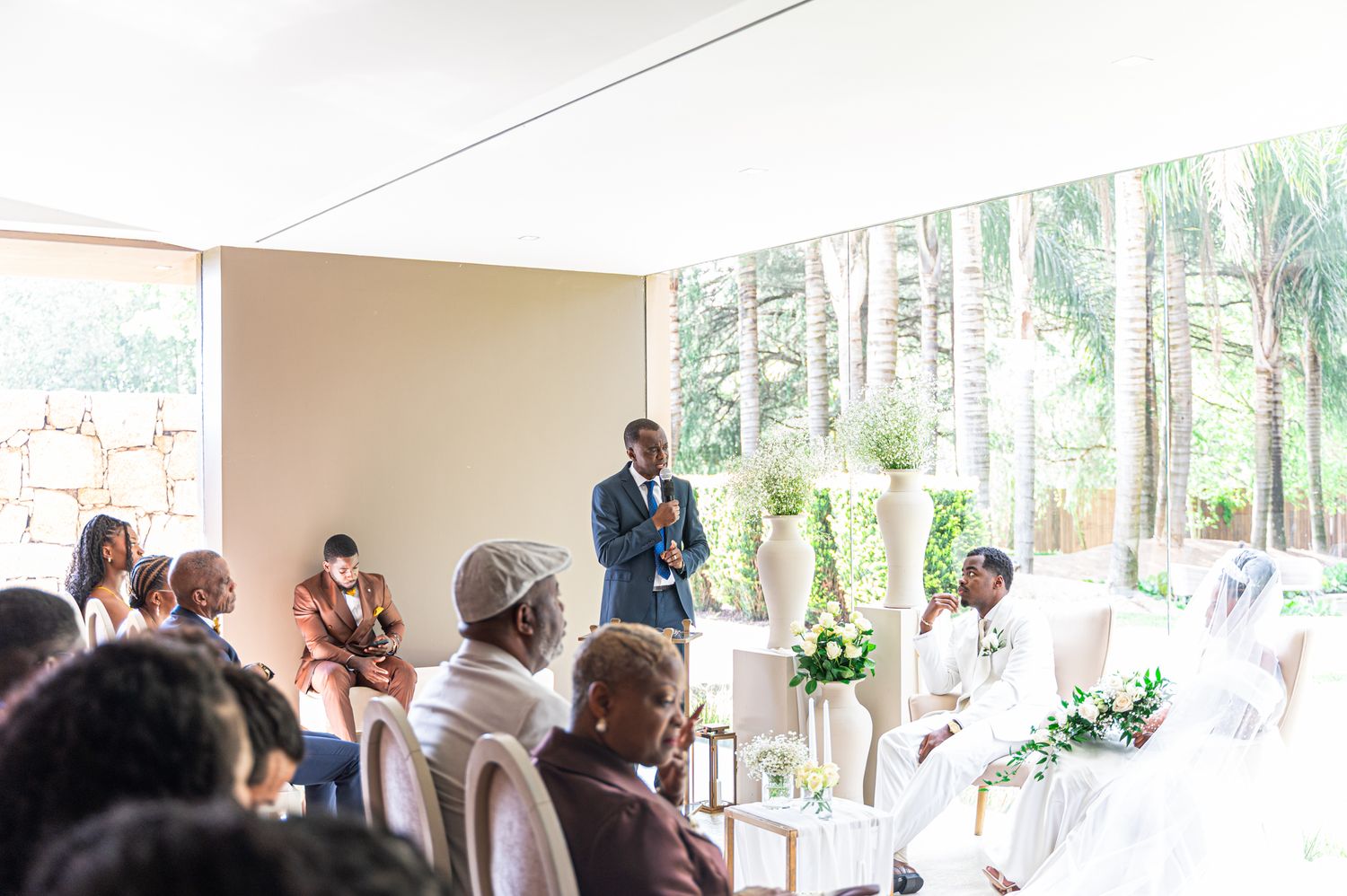 Wedding officiant in blue suit addresses seated guests in a bright modern ceremony venue.